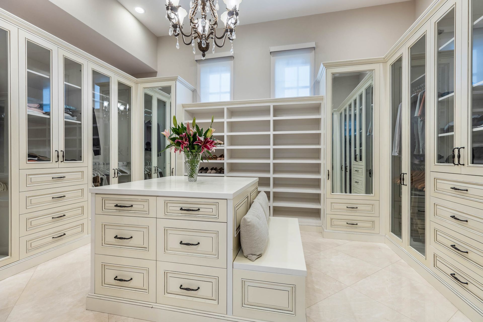 Luxurious white walk-in closet with island, shelves, and glass-door cabinets, under chandelier.