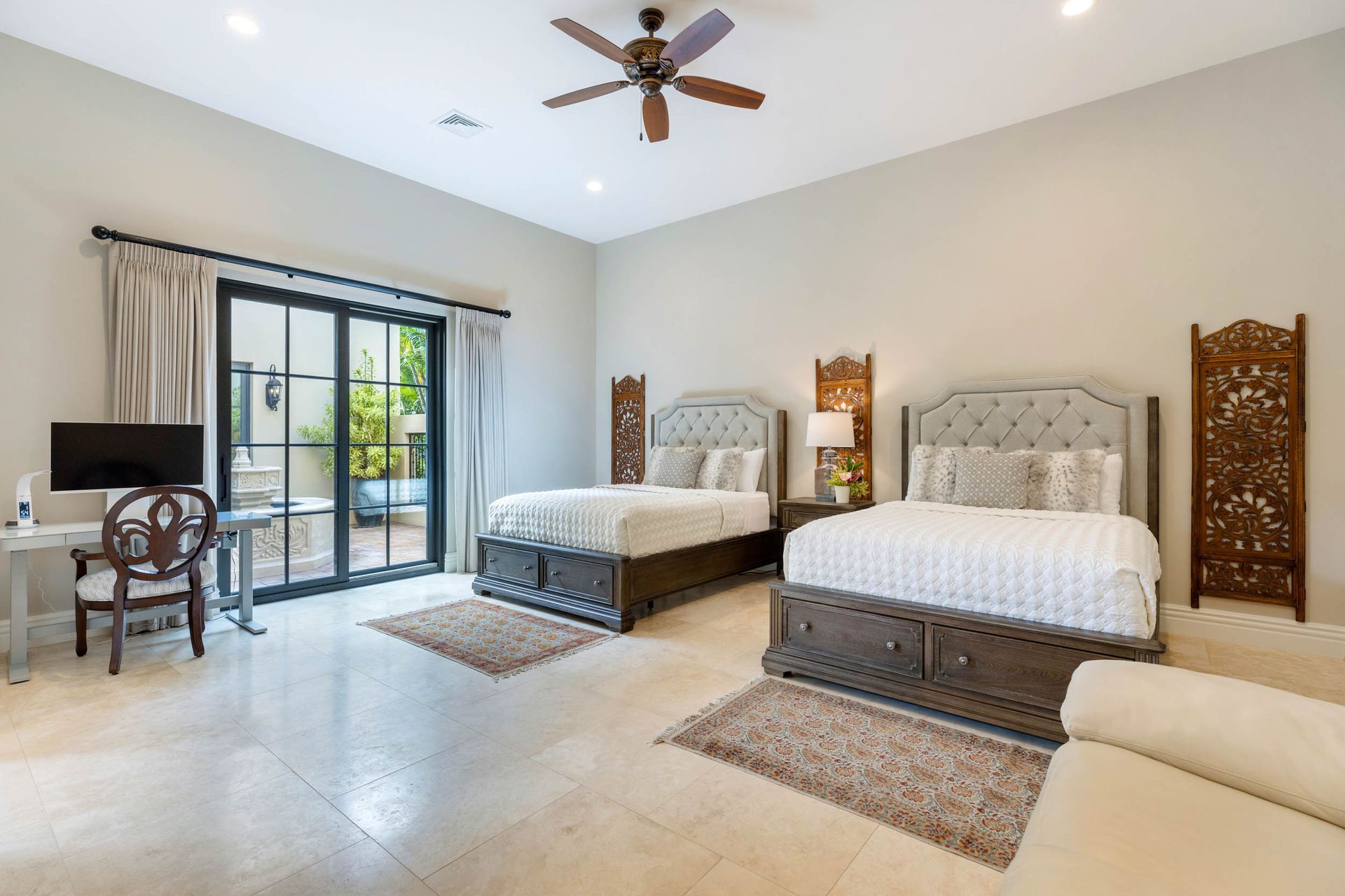 Spacious bedroom with two beds, desk, and sliding door to a patio. Beige walls, light flooring, and decorative screens.