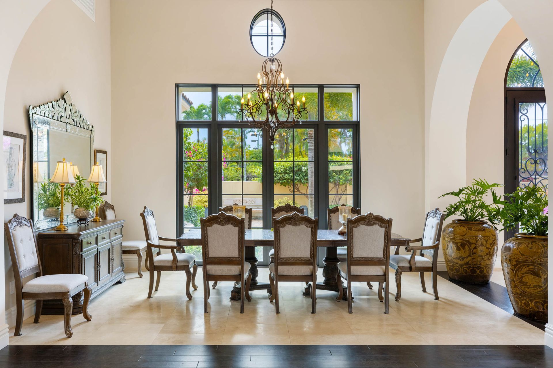 Elegant dining room with large table, chairs, chandelier, and a large window overlooking greenery.