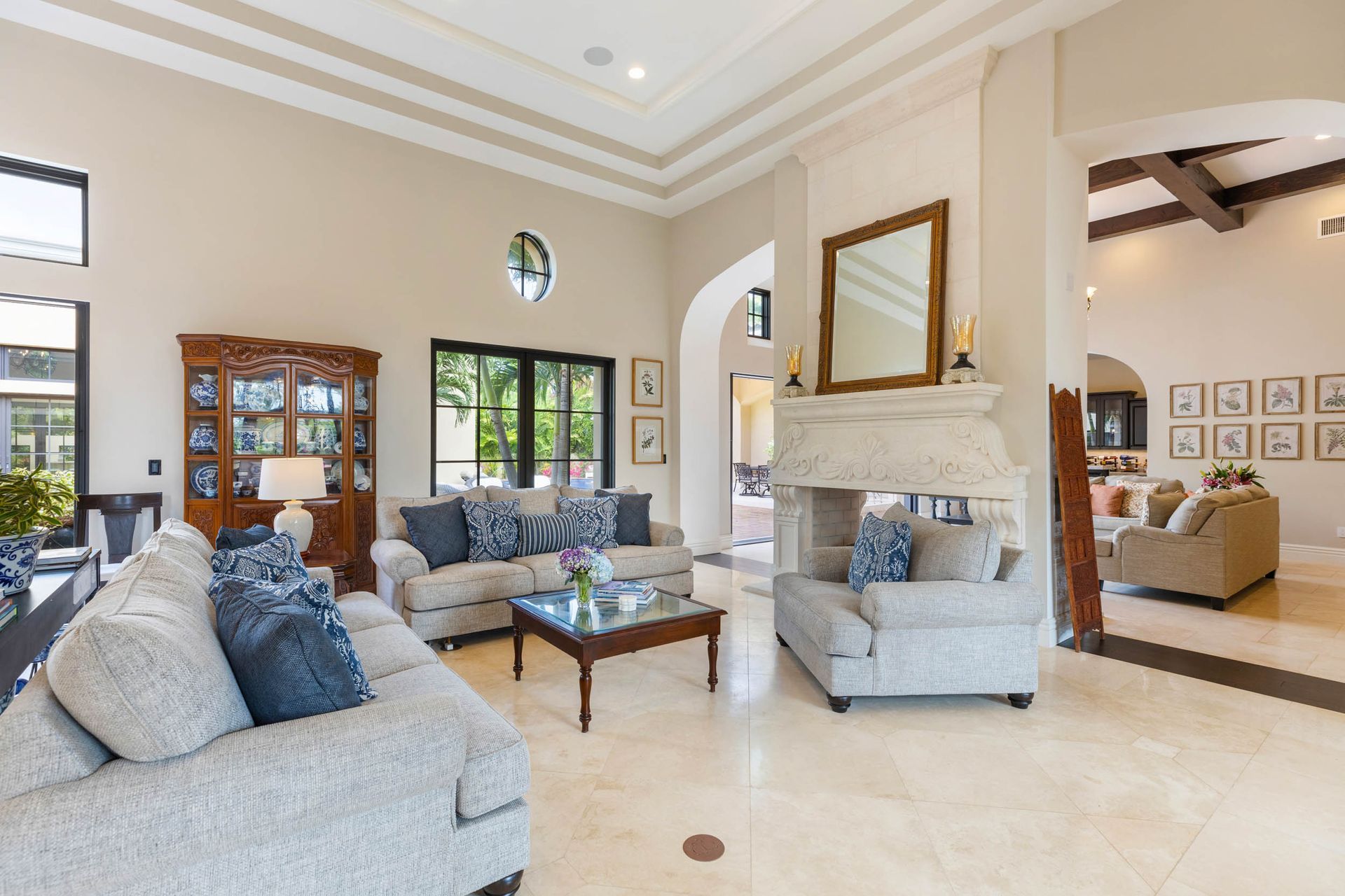 Living room with light-colored walls, marble floors, and three sofas. Sunlight streams through a window.