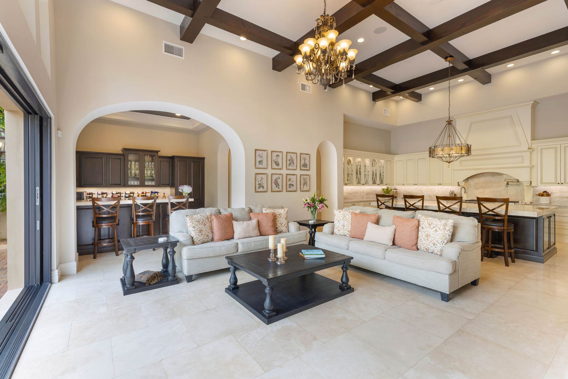 Spacious living room with sofas, kitchen, and arched doorways. Cream walls, wood beams, and large windows.