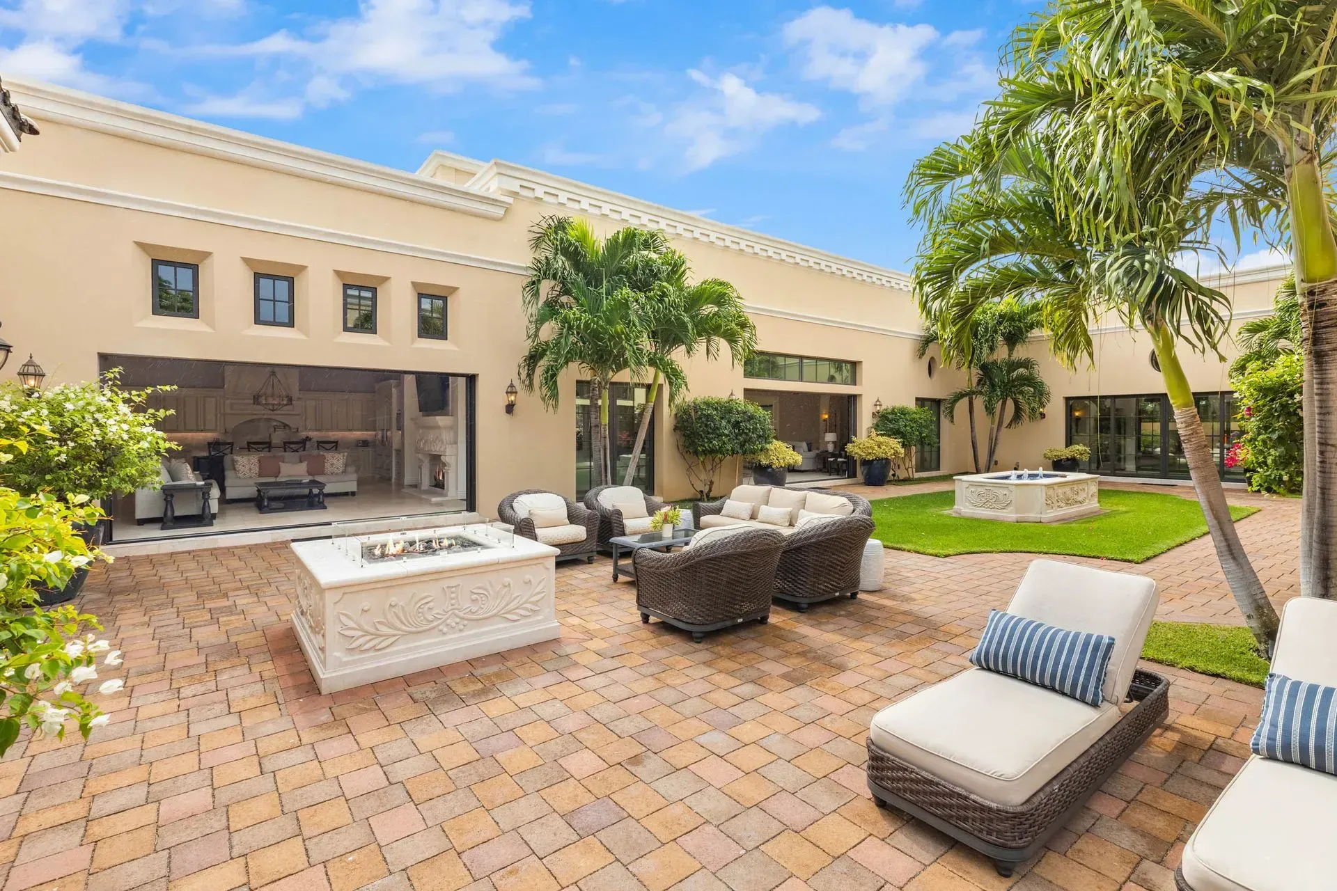 Outdoor patio with seating, fire pit, and lawn, surrounded by a beige building with open doors and palm trees.