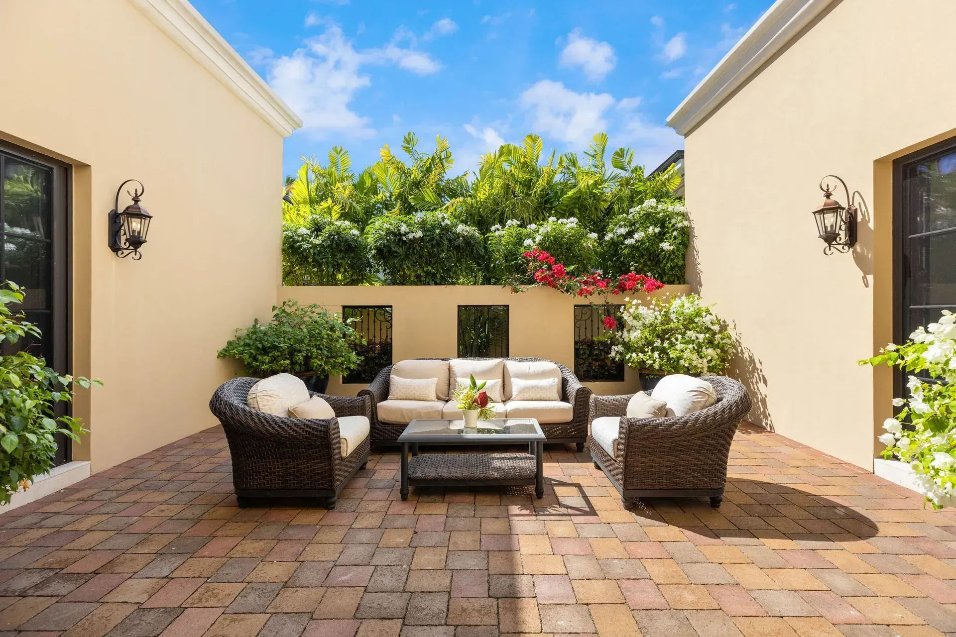 Patio with outdoor furniture, brick floor, surrounded by walls and greenery under a blue sky.