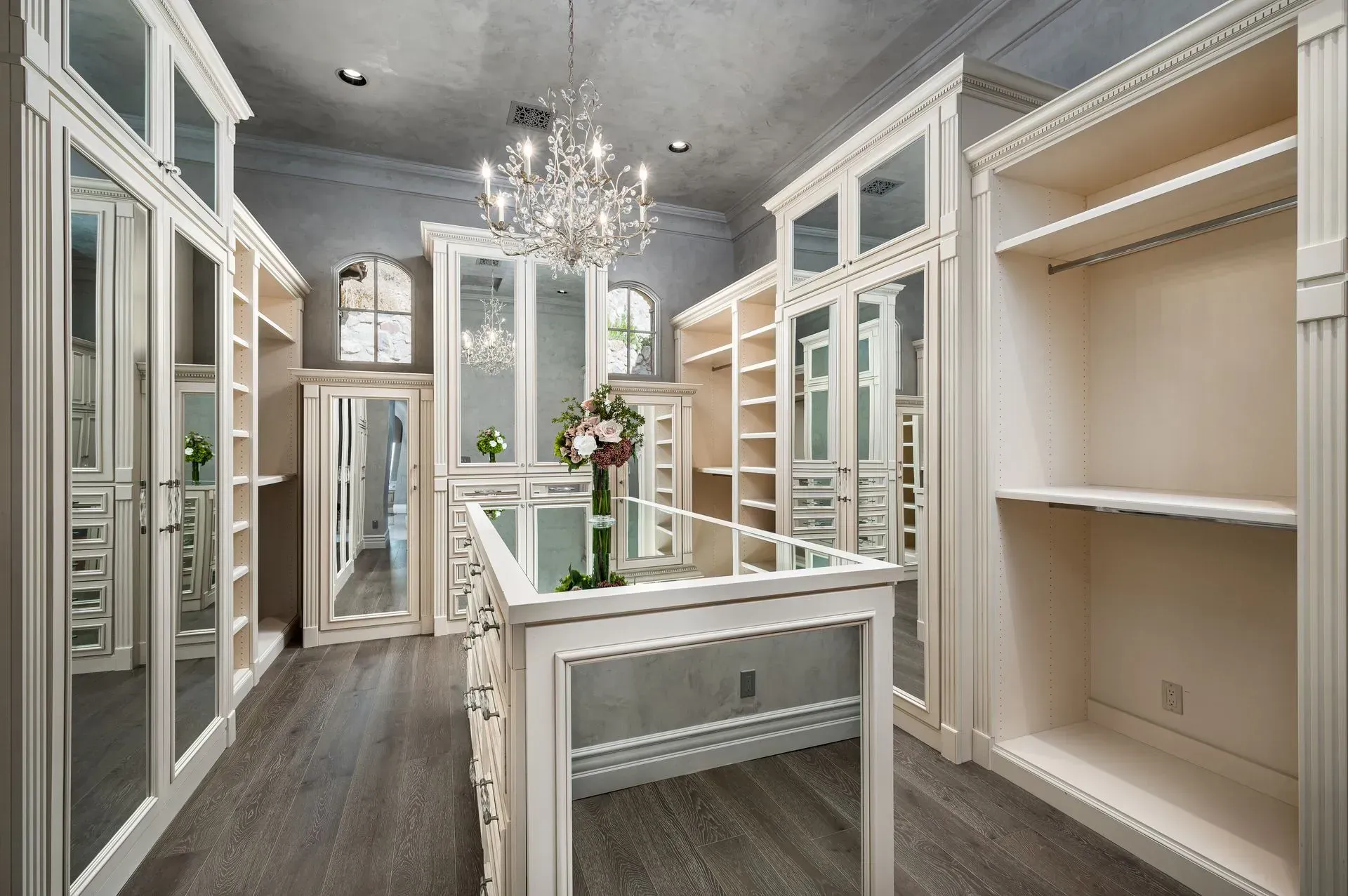 Elegant white walk-in closet with mirrored doors, shelves, and a chandelier; gray flooring and ceiling.