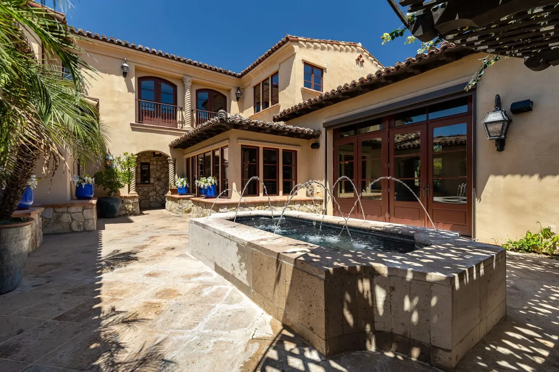 Courtyard with fountain, two-story beige house, brown doors, and stone pathway under a sunny sky.