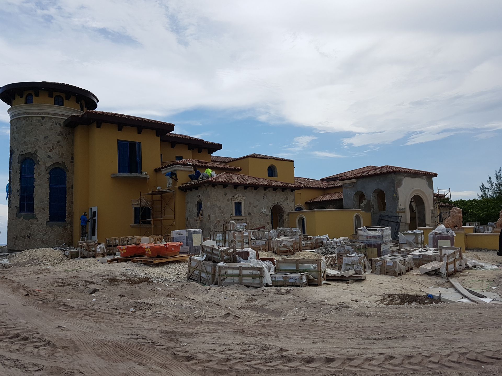 Construction site of a yellow stucco Mediterranean-style house with stone accents, blue windows, and a cloudy sky.