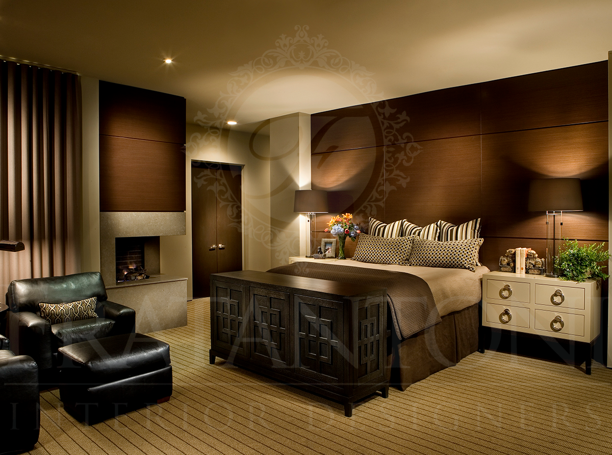 A modern hotel bedroom with a large bed, leather armchair, stone fireplace, and dark wood accents in warm, neutral tones.