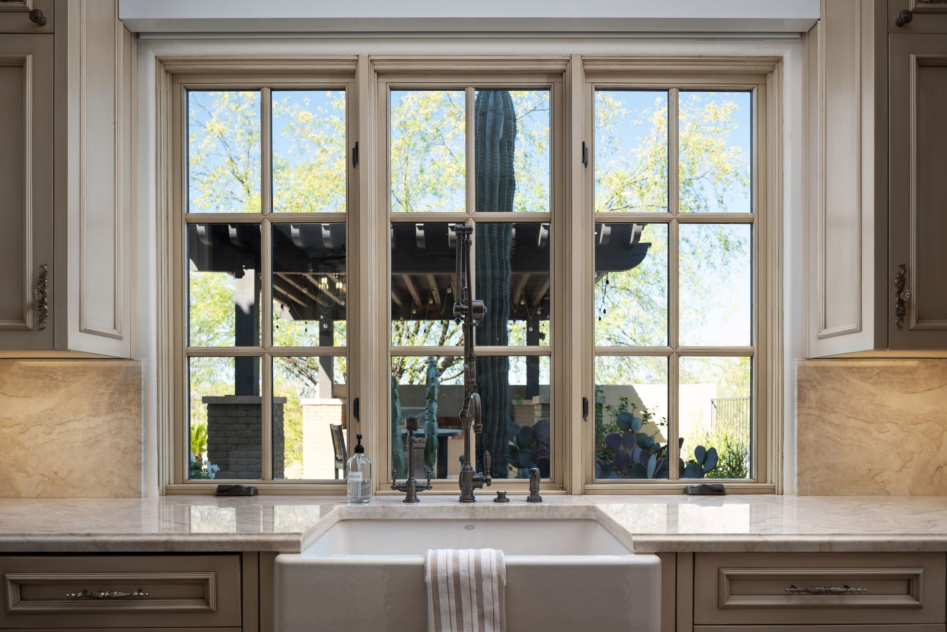 Kitchen sink with three-pane window, view of outdoor pergola. Beige cabinets, white sink, and light countertop.
