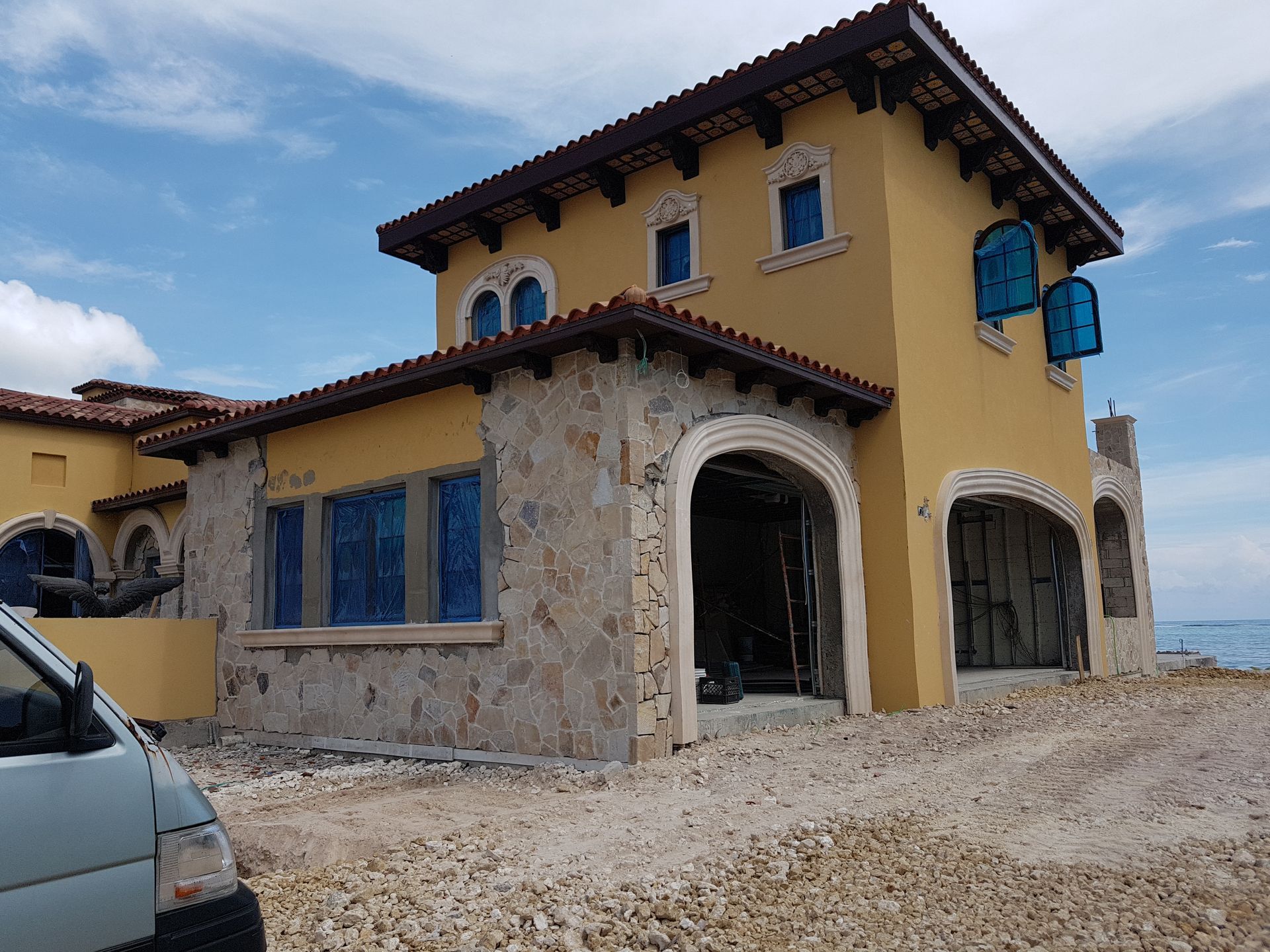 Yellow stucco and stone house with blue window covers under a blue sky, near a body of water.