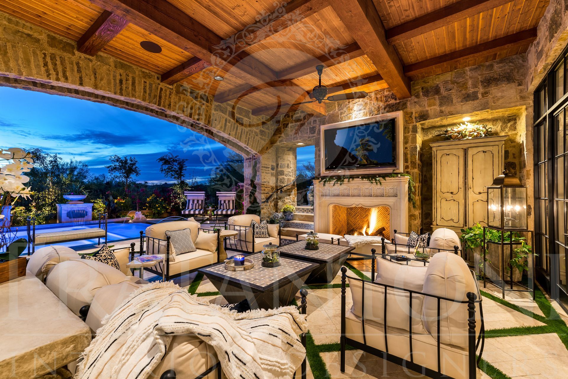 Elegant covered outdoor patio with fireplace, seating, mounted tv, a brick arch, and pool view at dusk.