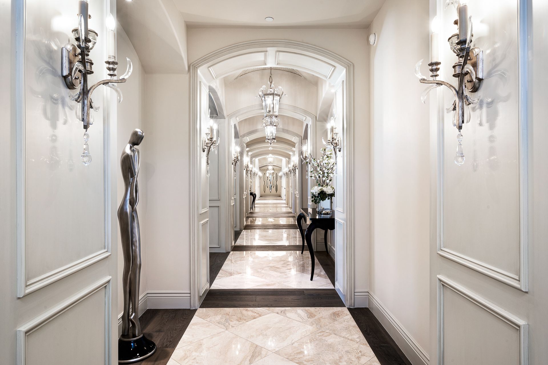 Long hallway with ornate white molding, sconces, and marble flooring. A statue stands near the entrance.