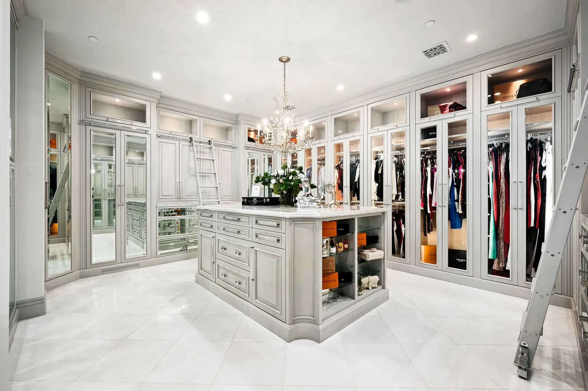 Spacious, elegant walk-in closet with central island and hanging clothes. White, reflective surfaces. Chandelier.