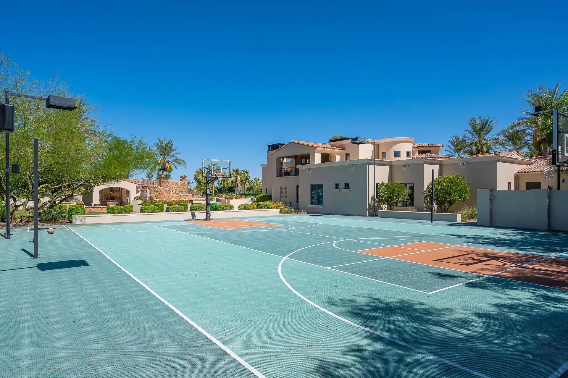 Outdoor basketball court with teal and brown surface, buildings in the background under blue sky.