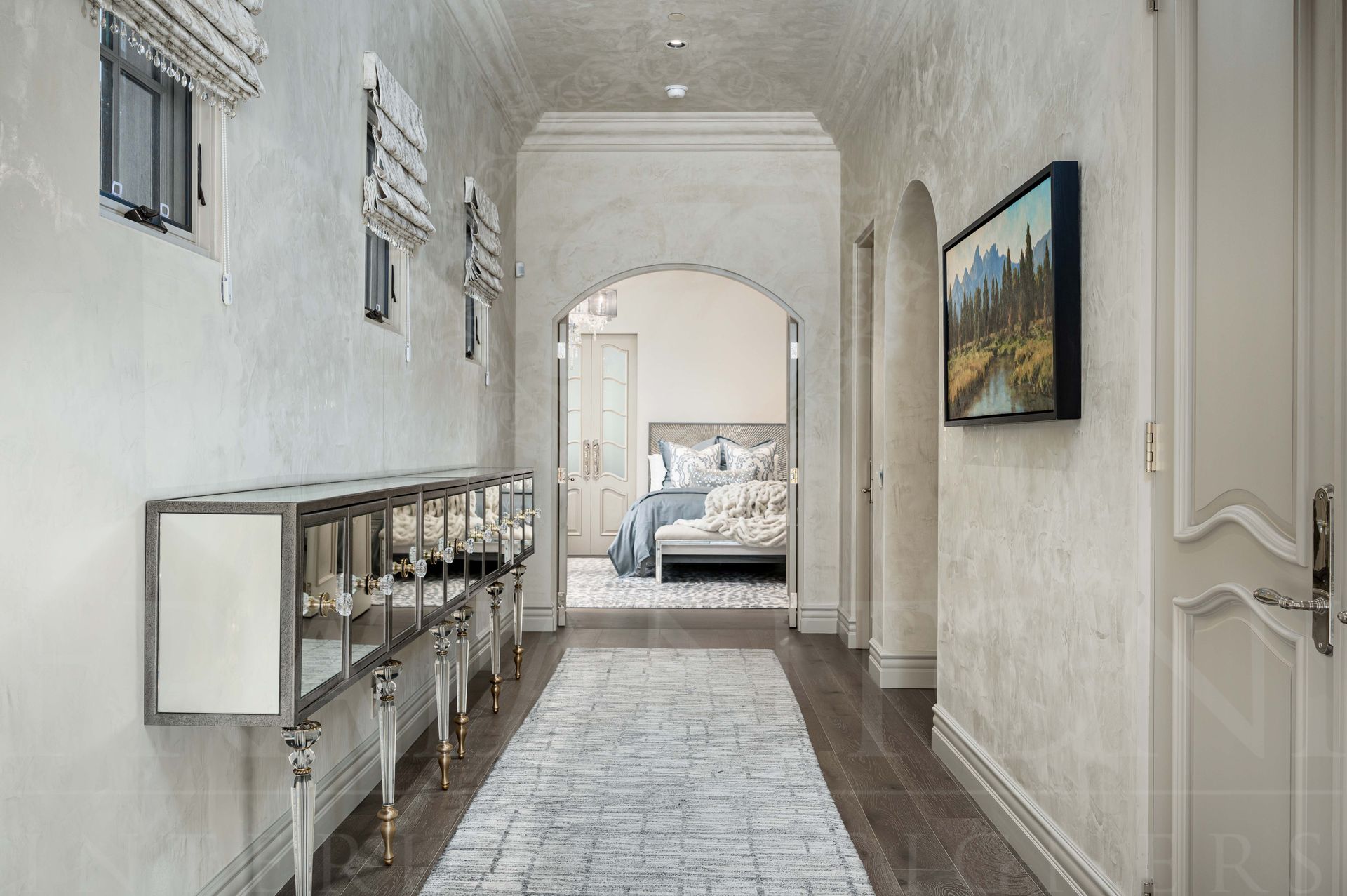 Bright hallway with lime washed walls, patterned runner, mirrored glass console table, and an arched view into a master bedroom.