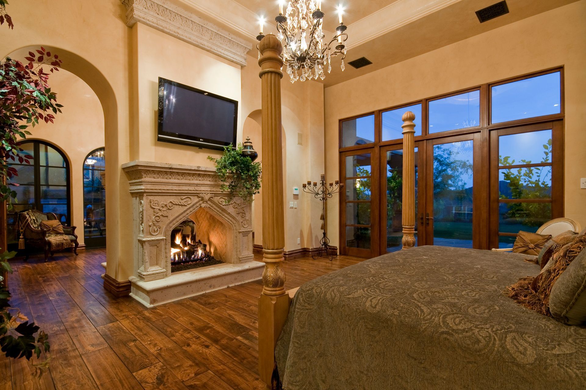 Luxurious bedroom with fireplace, TV, and large windows overlooking a yard.