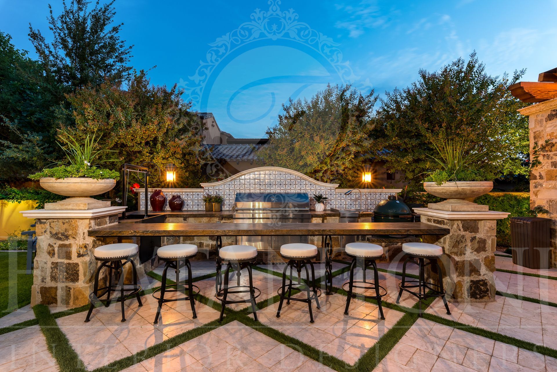 Outdoor kitchen with bar seating, grill, and lush desert landscaping at twilight.