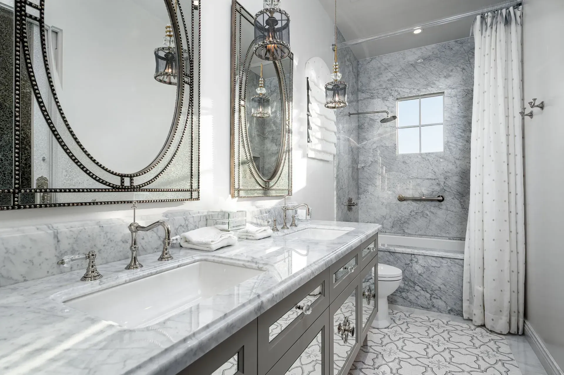 Luxurious white and gray bathroom with marble countertops, mirrored vanities, and ornate light fixtures.
