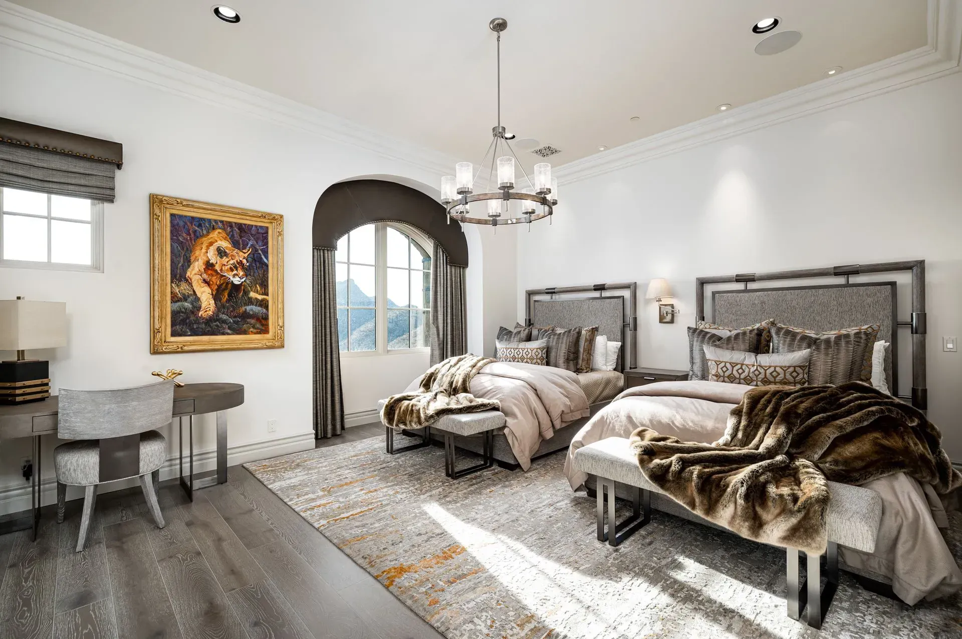 Elegant bedroom with two beds, artwork, desk, and arched window overlooking a mountain view.