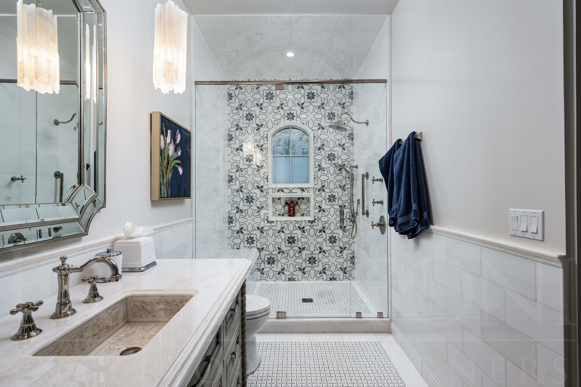Bright white bathroom with grey ornate vanity, large mirror, patterned mosaic shower wall, marble inlaid sink, and dark blue towels