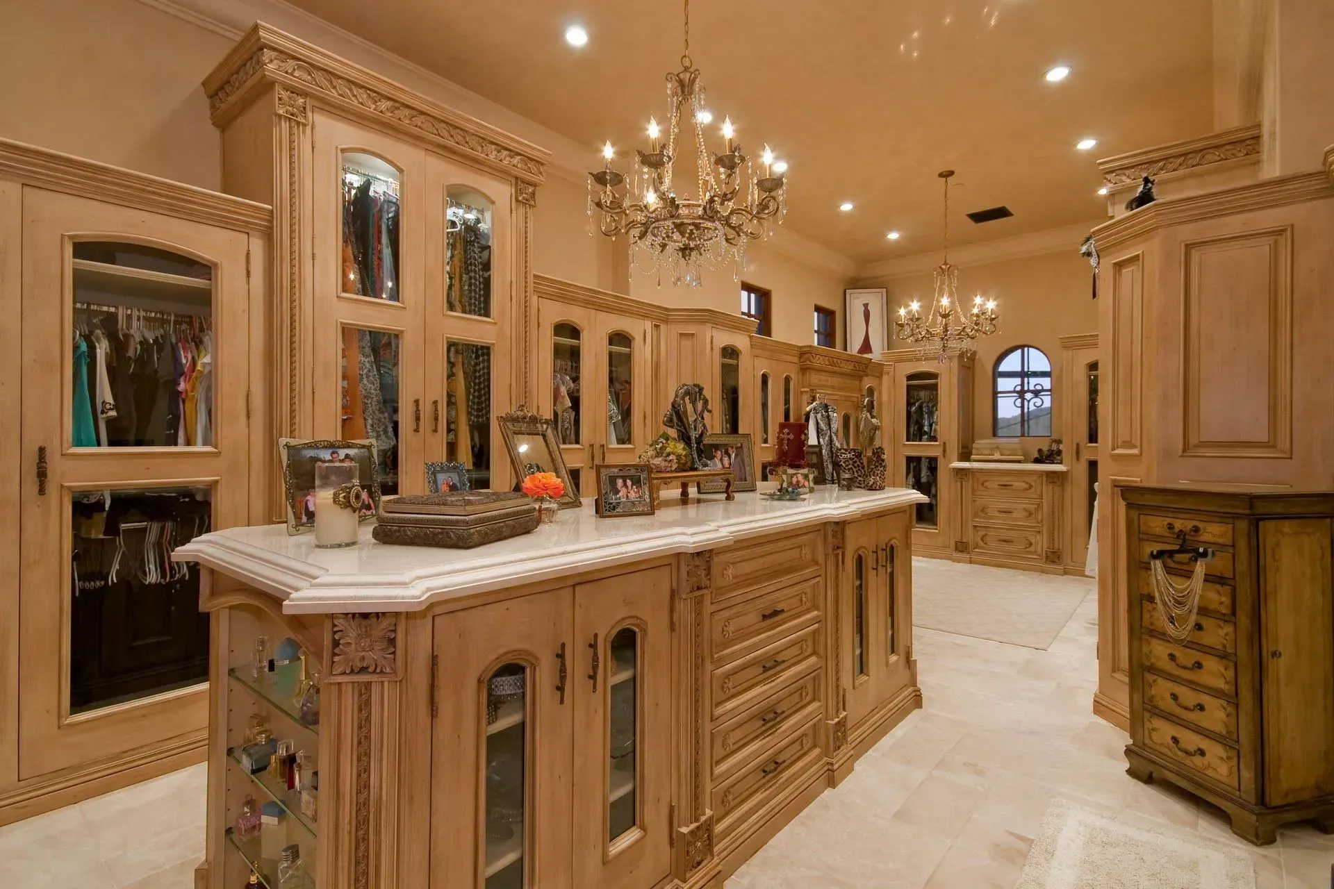Luxurious, ornate walk-in closet with custom light wood cabinetry, marble countertop island, and crystal chandeliers.