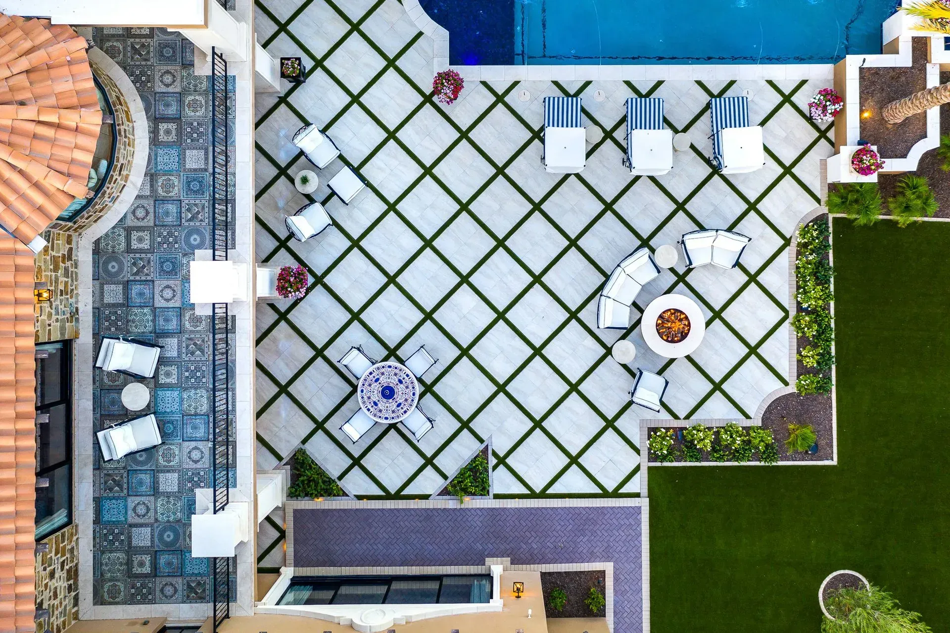 Aerial view of a patio with a swimming pool, lounge chairs, seating areas, and a fire pit. Green grass, blue accents.