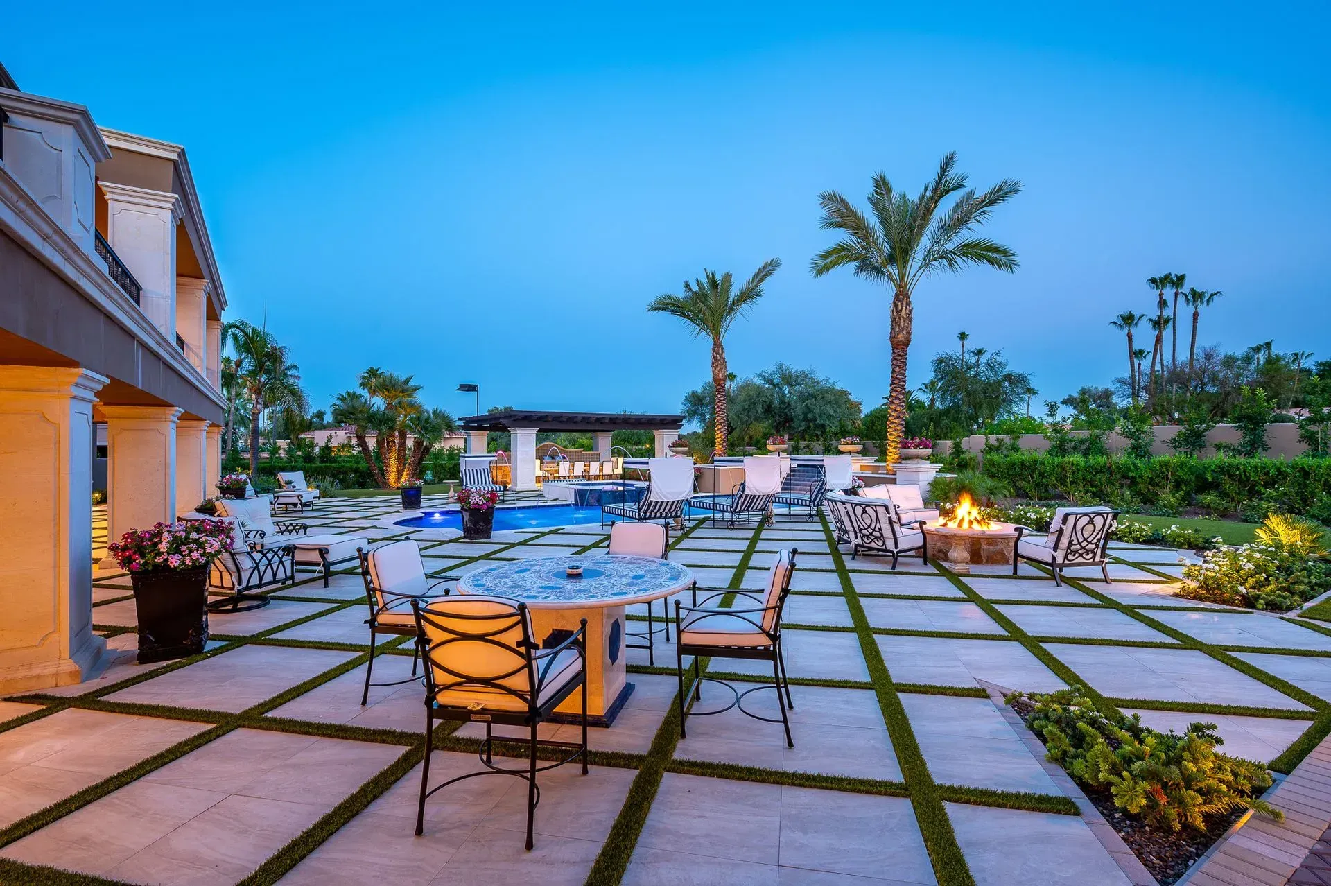 Patio with seating around a fire pit and dining table, with a pool, palm trees, and a house in the background.