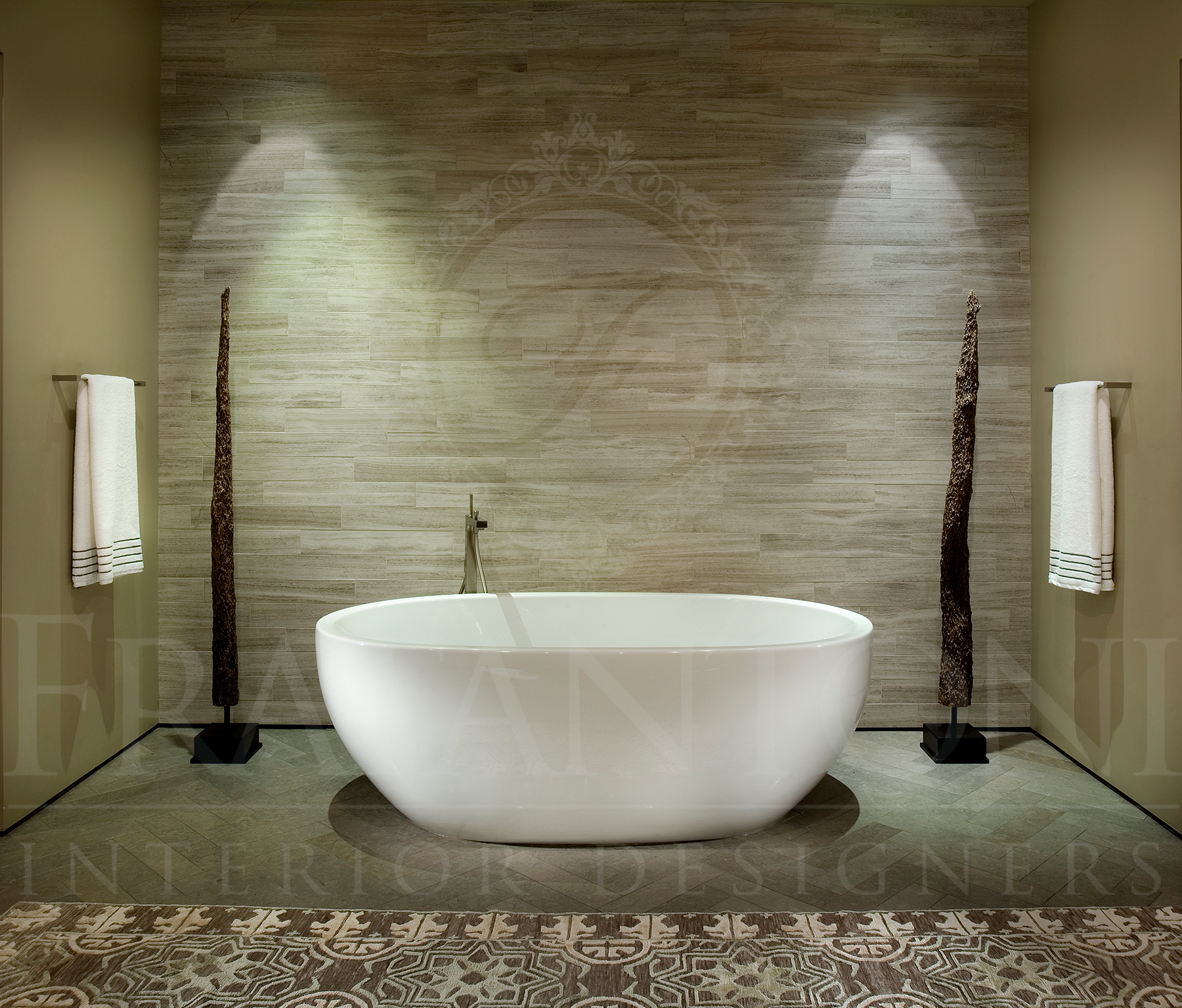 A white freestanding bathtub sits centered against a textured stone accent wall with two spotlights and floor sculptures.