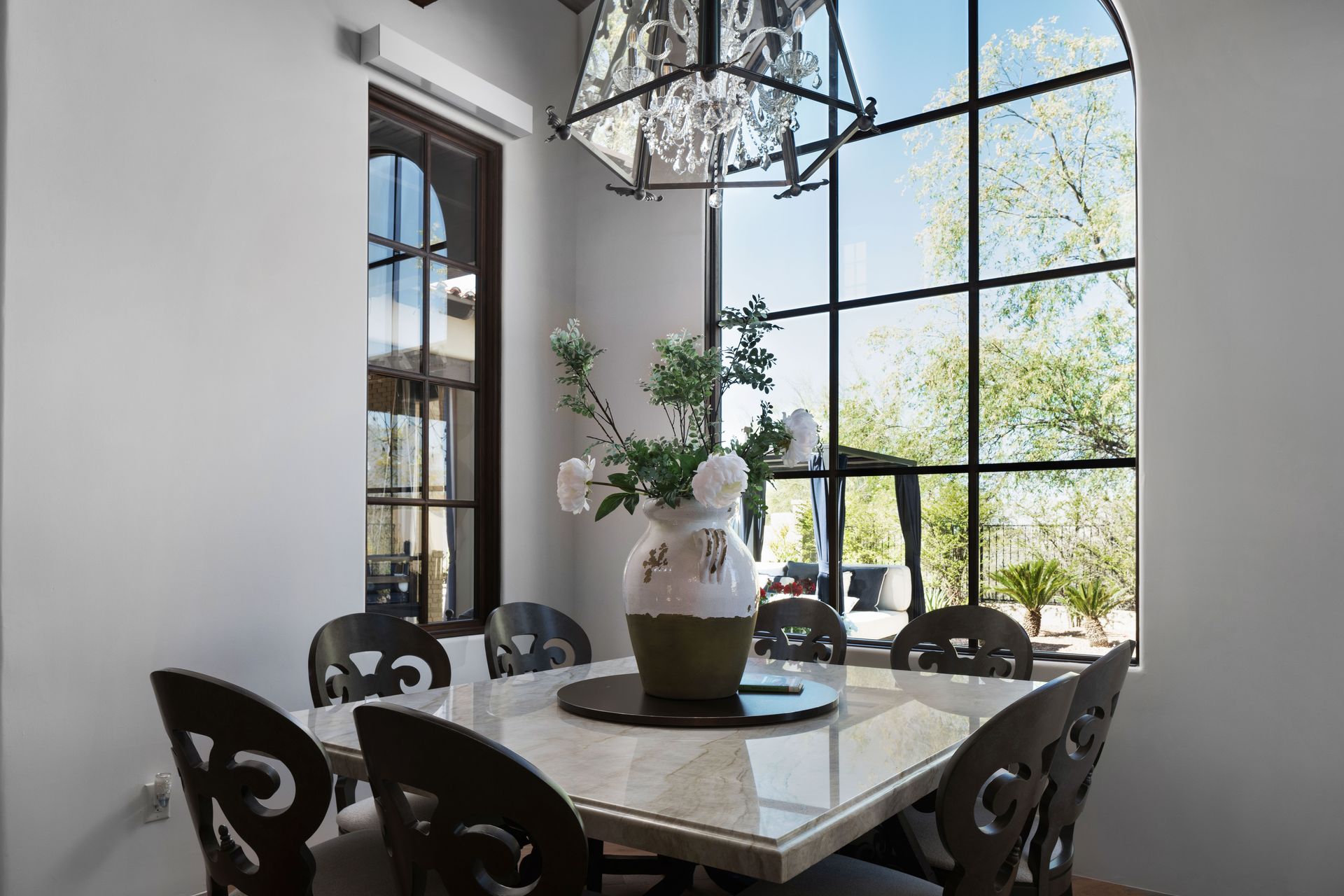 Dining room with marble table, ornate chairs, large arched window, chandelier, and a vase of flowers.