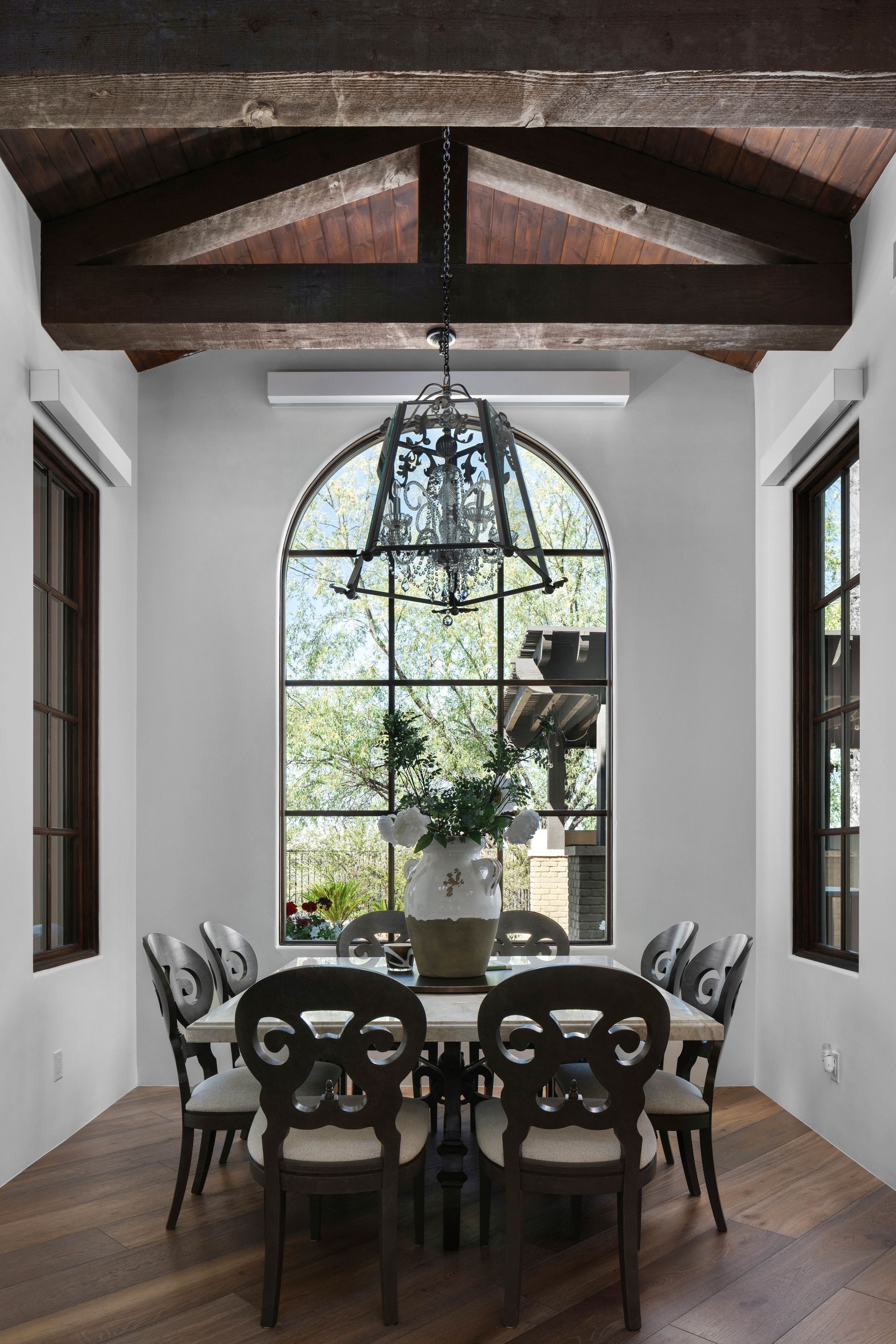 Dining room with arched window, chandelier, dark wood beams, and a table set for a meal.