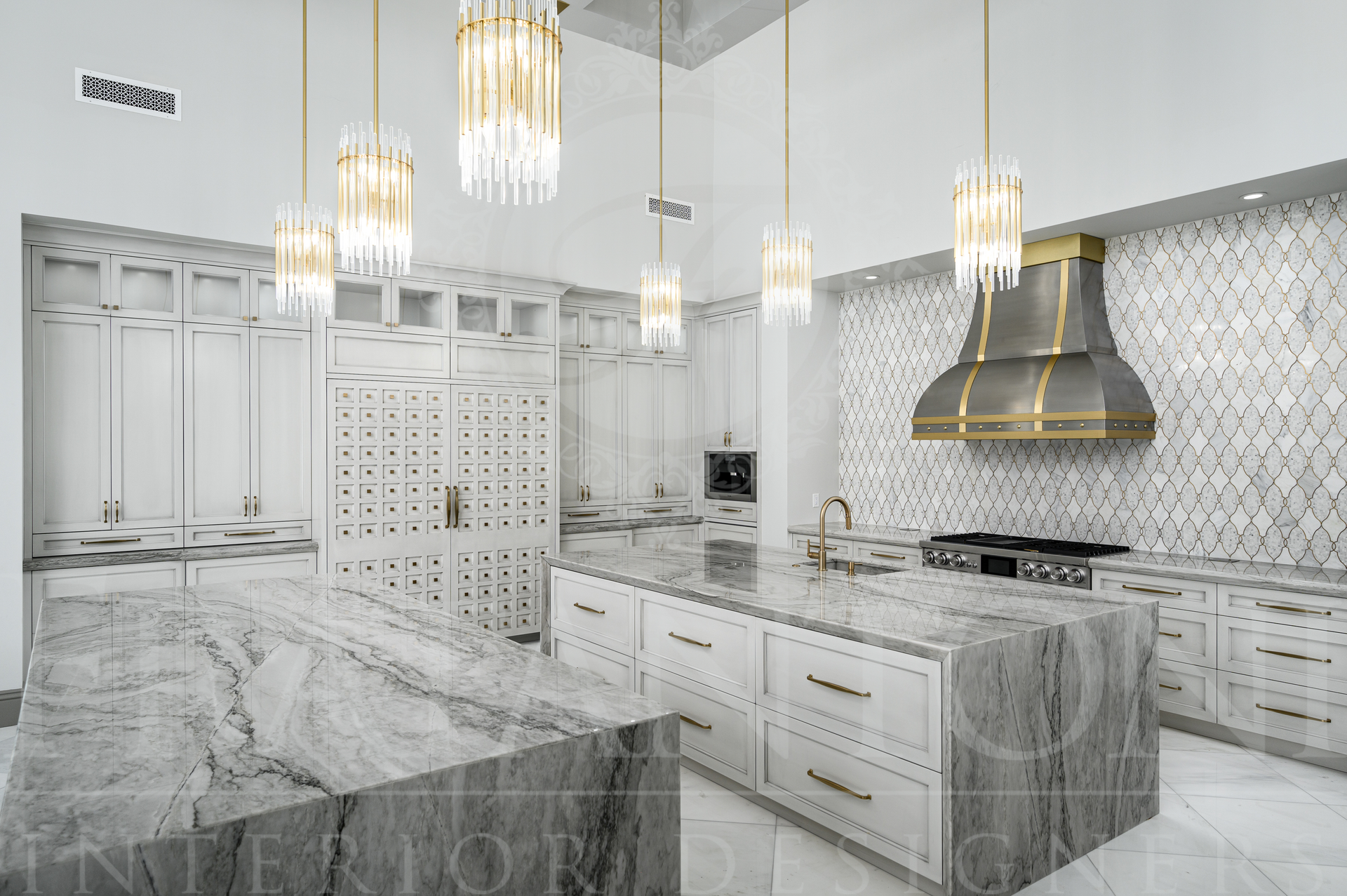 Luxury kitchen with double marble islands, white cabinets, gold chandeliers, and patterned backsplash.
