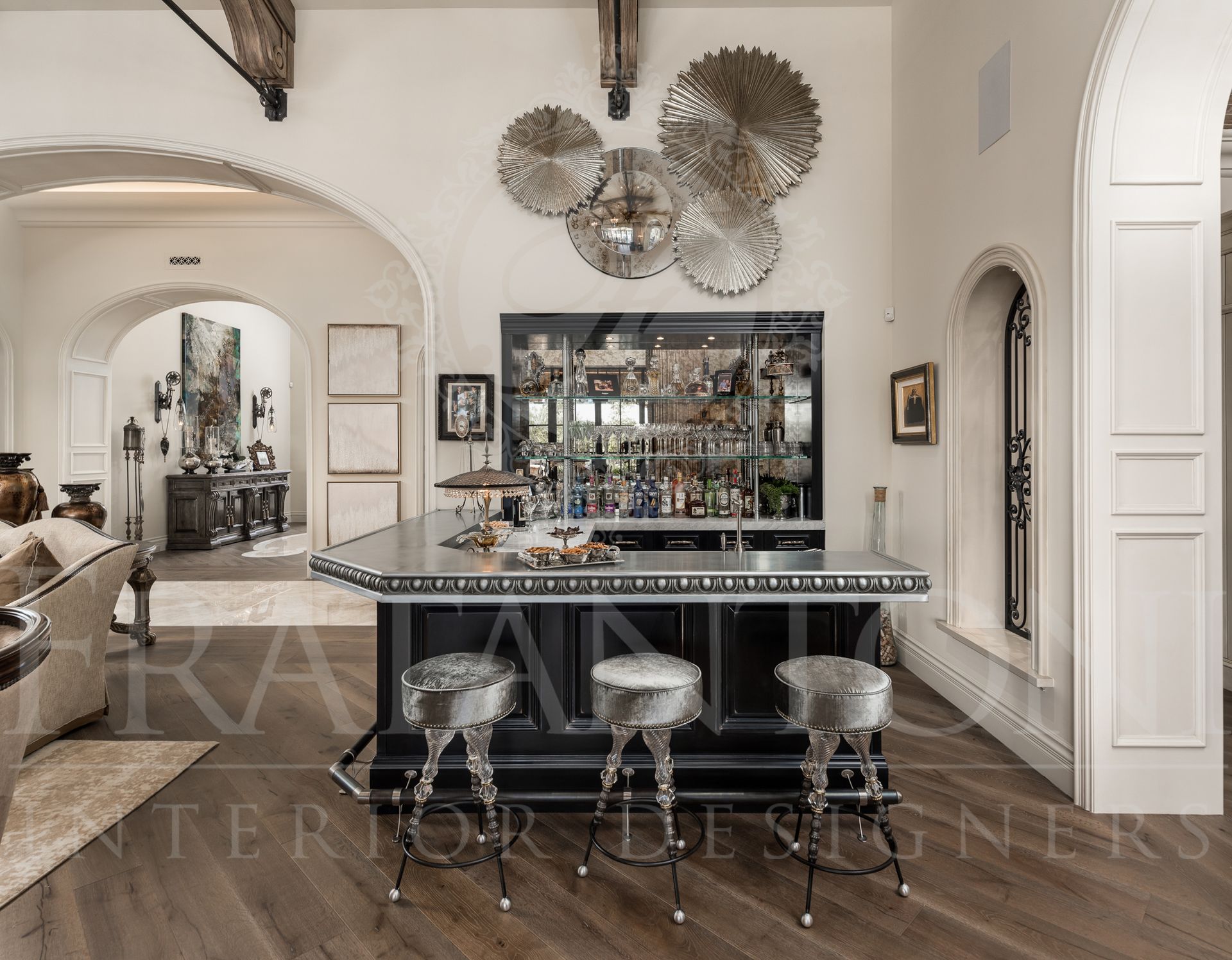 Custom home bar with stools, mirrored backdrop, and metal art, in a room with arched doorways and wood flooring.