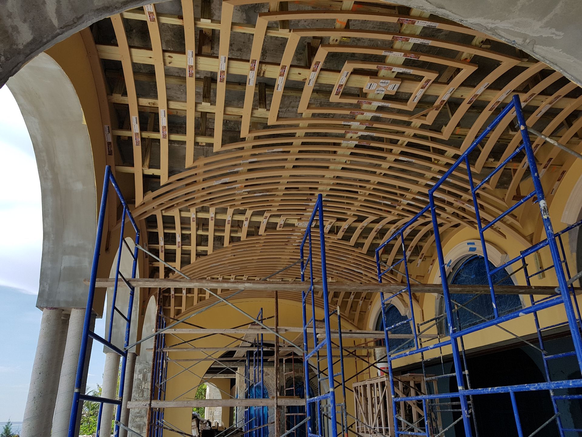 Construction of arched ceiling with wooden framework, scaffolding, and columns. Yellow walls, blue sky.
