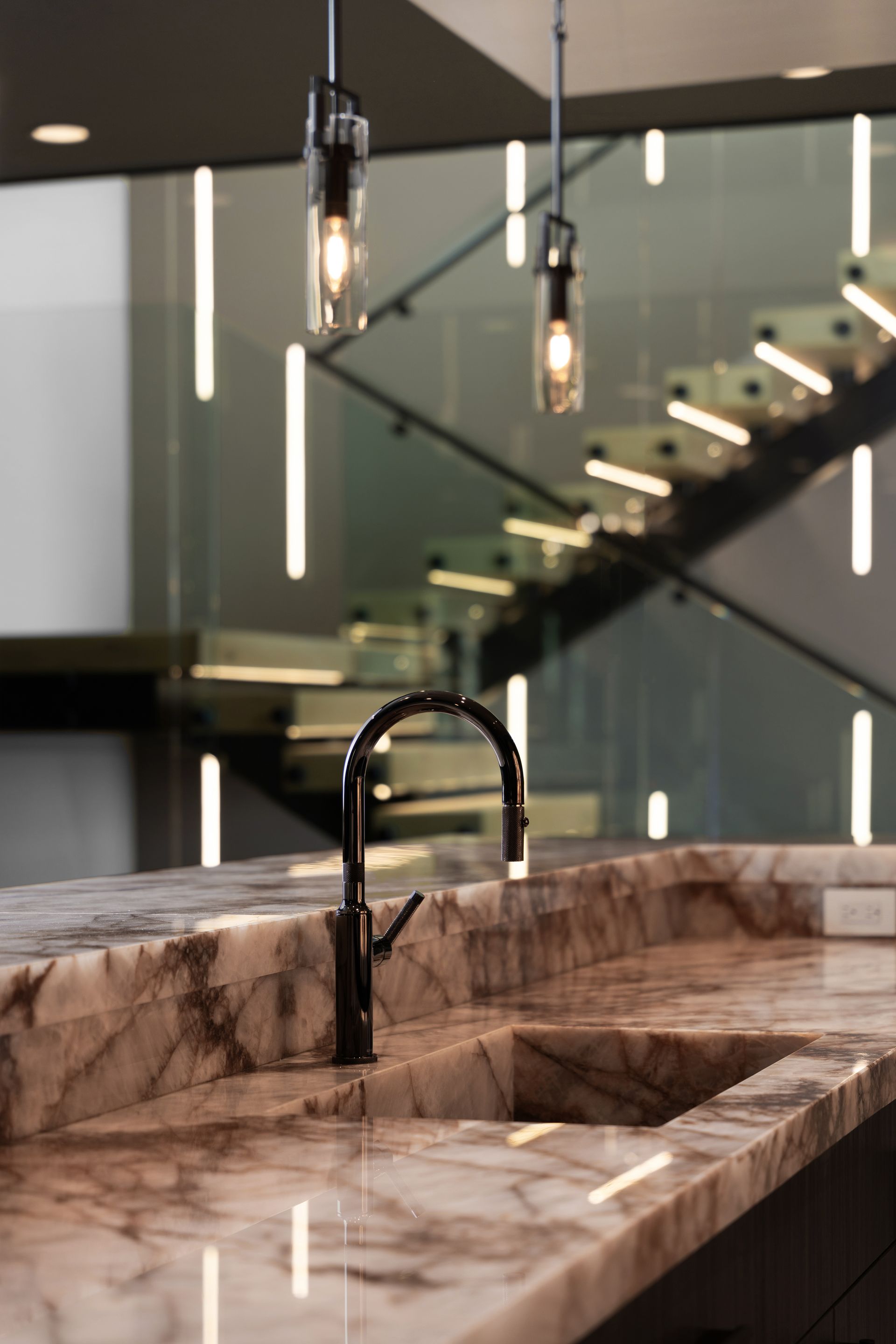 Marble countertop with a black faucet, lit pendant lights, and a staircase in the background.