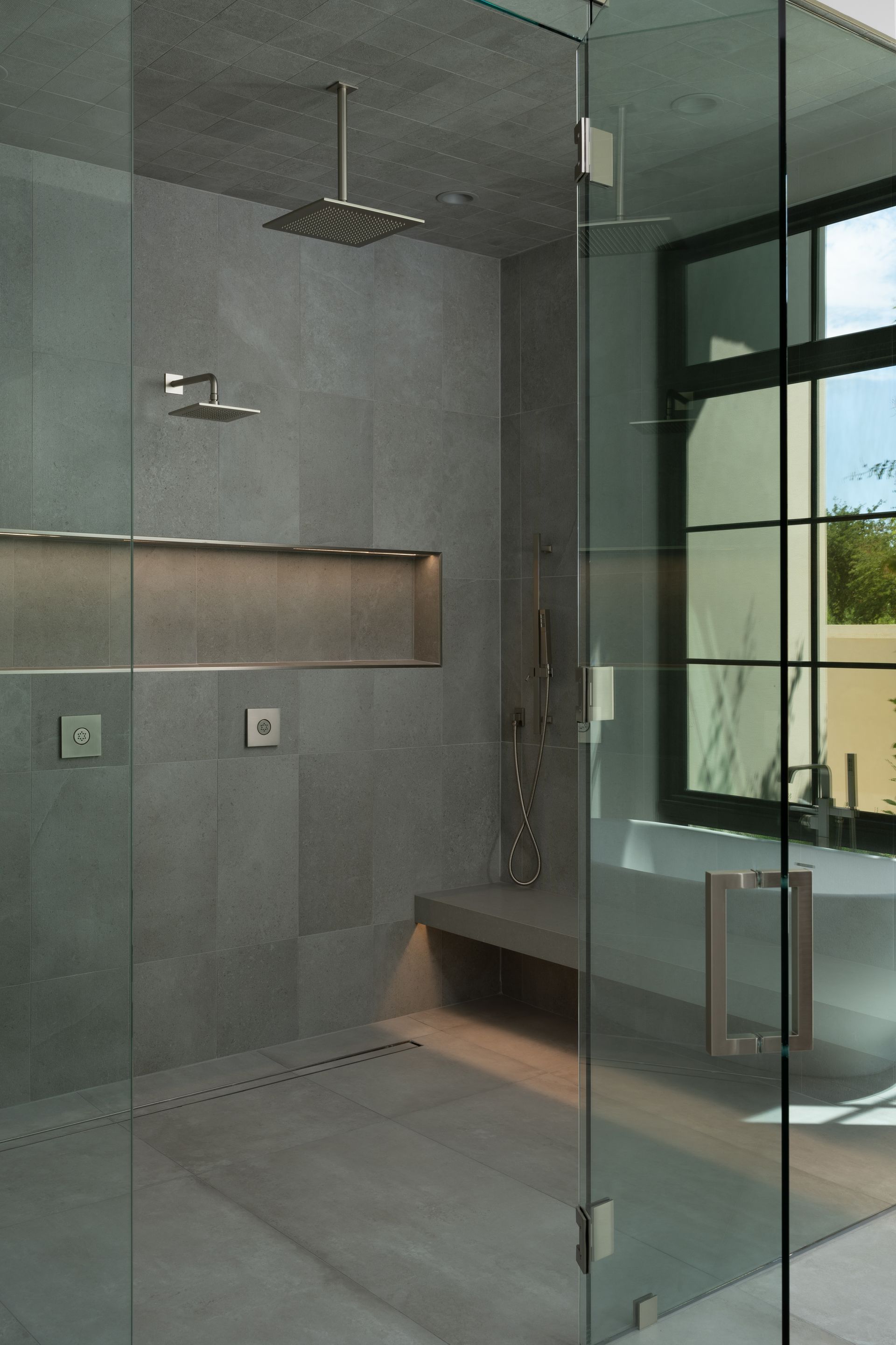 Modern gray-tiled shower with glass door, bench, and various showerheads; adjacent to a window and soaking tub.