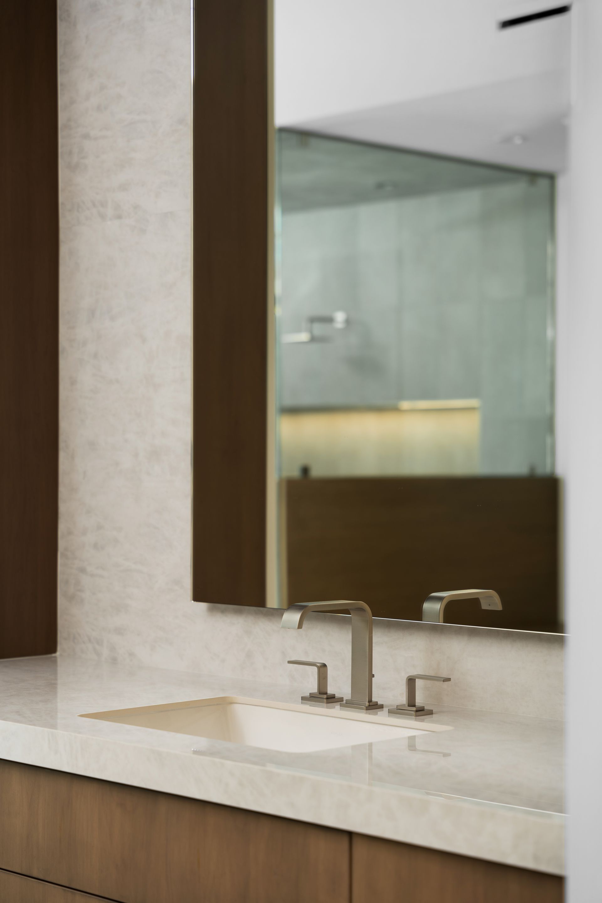 Bathroom with a rectangular mirror reflecting a shower. Beige countertop with sink and faucet. Brown cabinets.