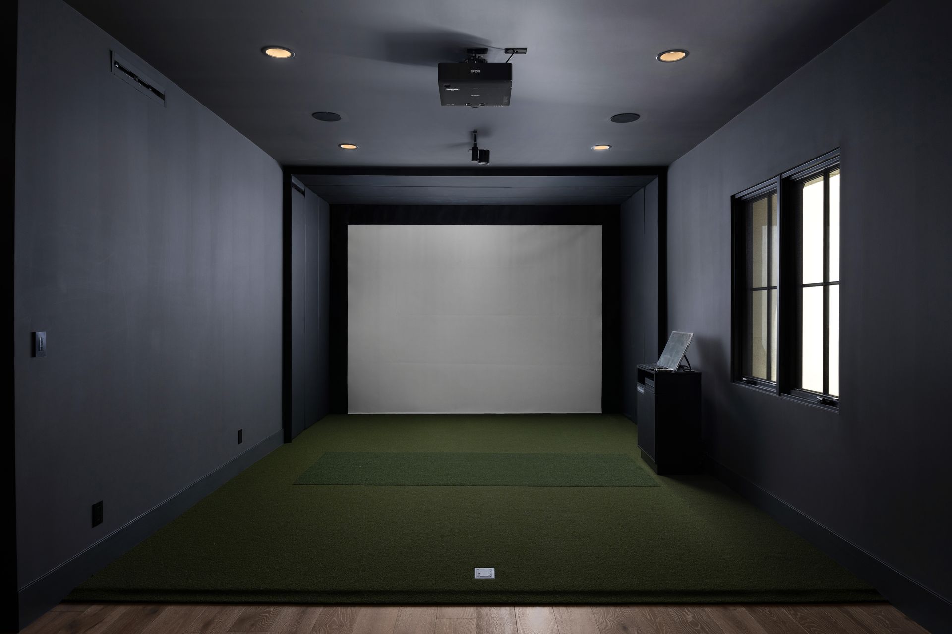 Dark gray home theater with projector, screen, and turf floor. Window on the right.