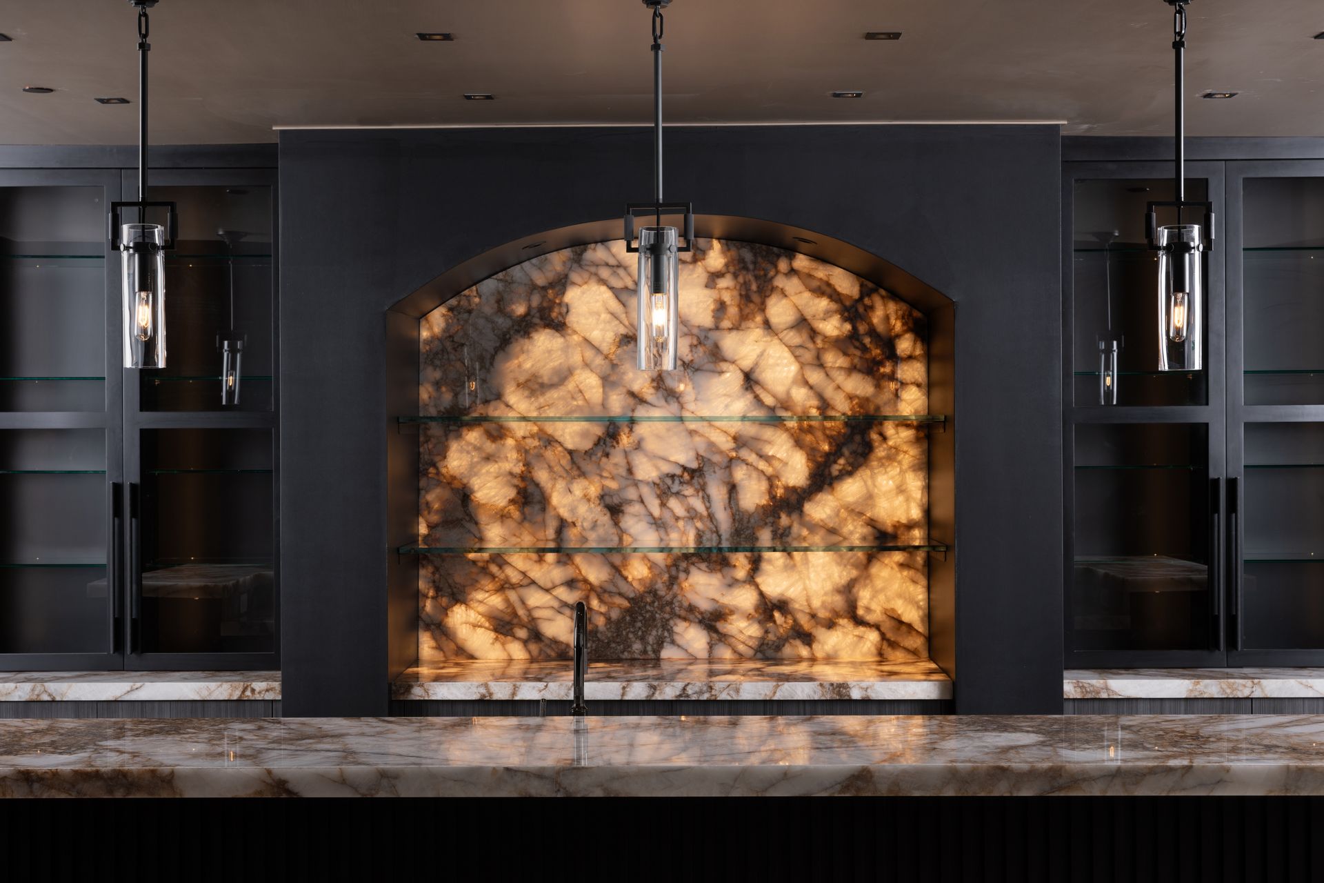 Bar with backlit, patterned wall, open shelving, and pendant lights. Dark cabinetry, marble countertop.