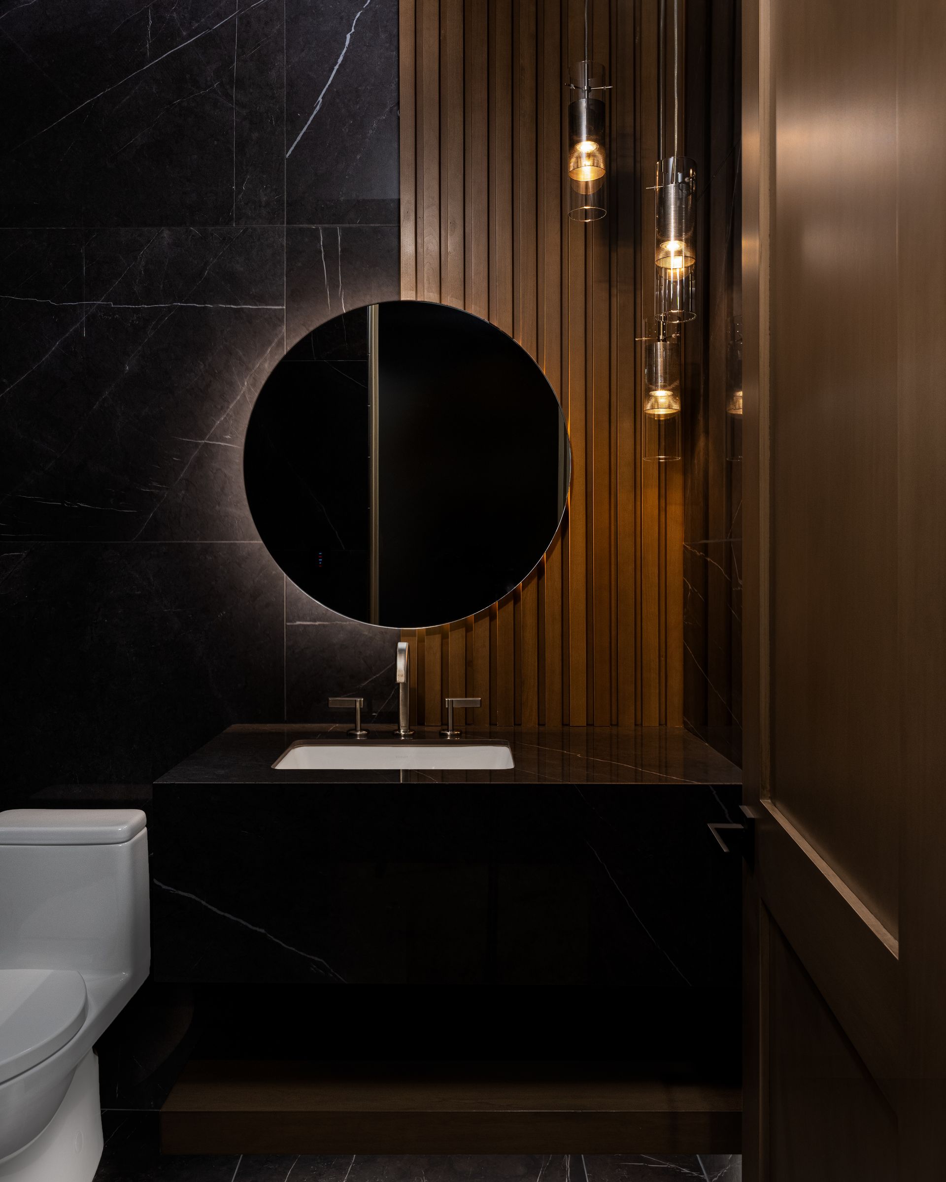 Stylish black bathroom with round mirror, black marble wall, wooden accents, and hanging lights.