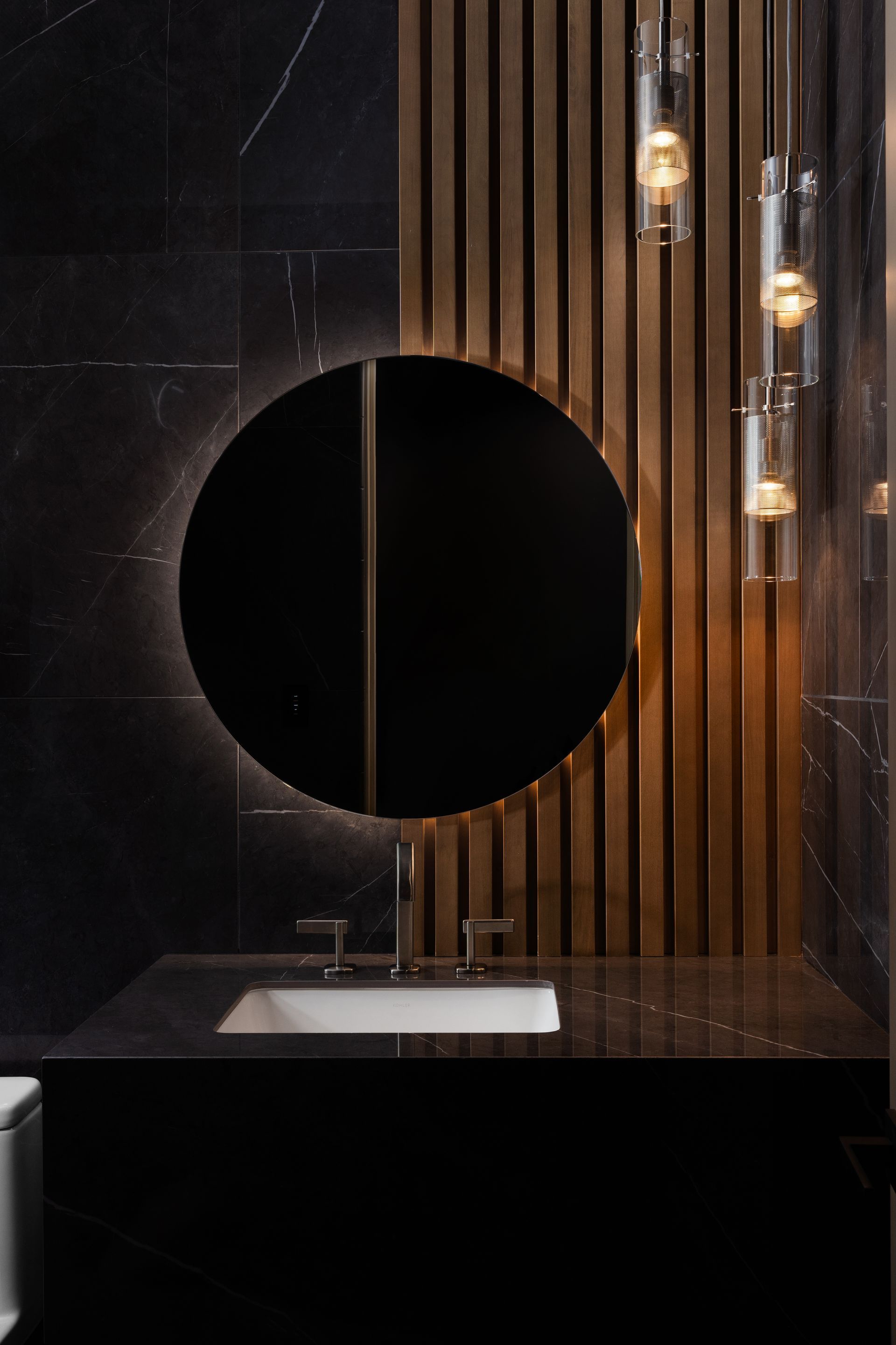 Bathroom with round mirror, black marble wall, wooden paneling, white sink, and hanging lights.