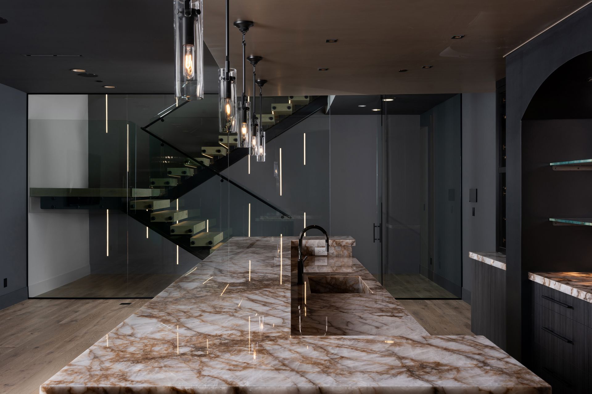 Modern kitchen with marble island, dark cabinets, floating staircase, and hanging lights.