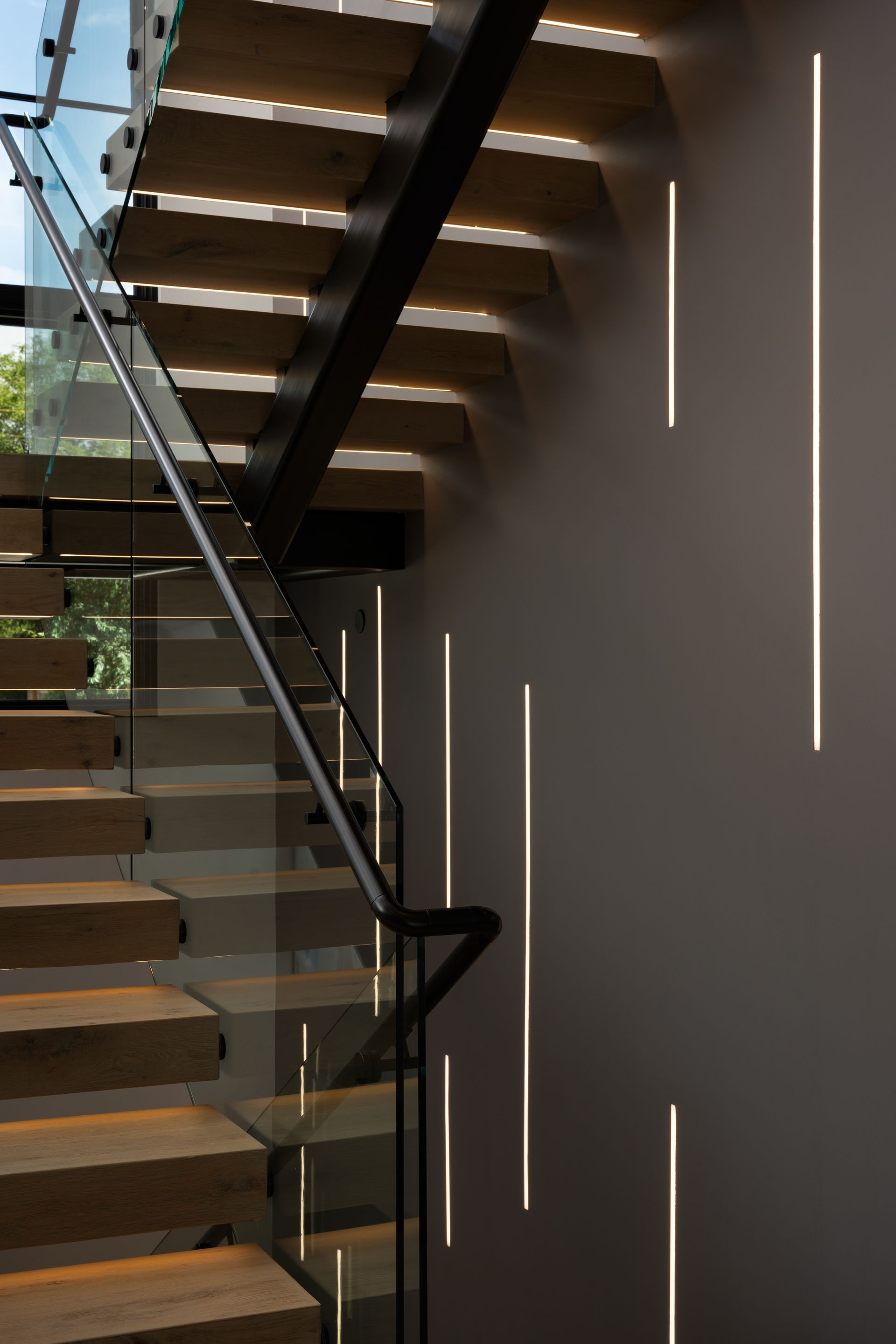 Staircase with wooden steps, a glass railing, and vertical wall lights casting shadows.