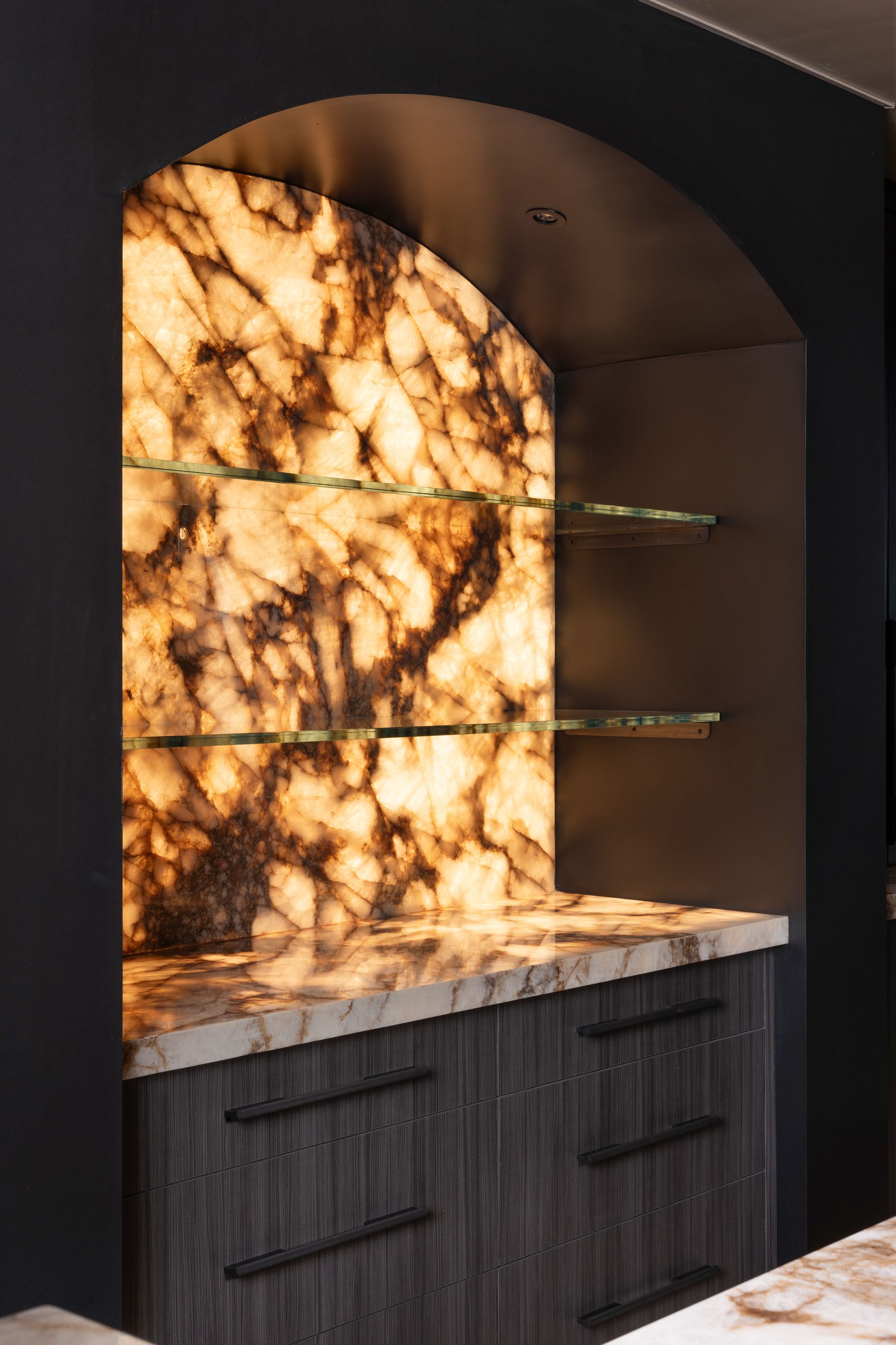 Lit onyx shelving niche with glass shelves and wood cabinet.