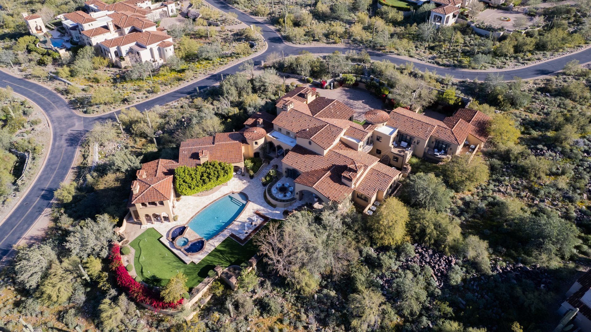 Aerial view of a large house with a pool, red tile roof, and a winding road in a desert landscape.