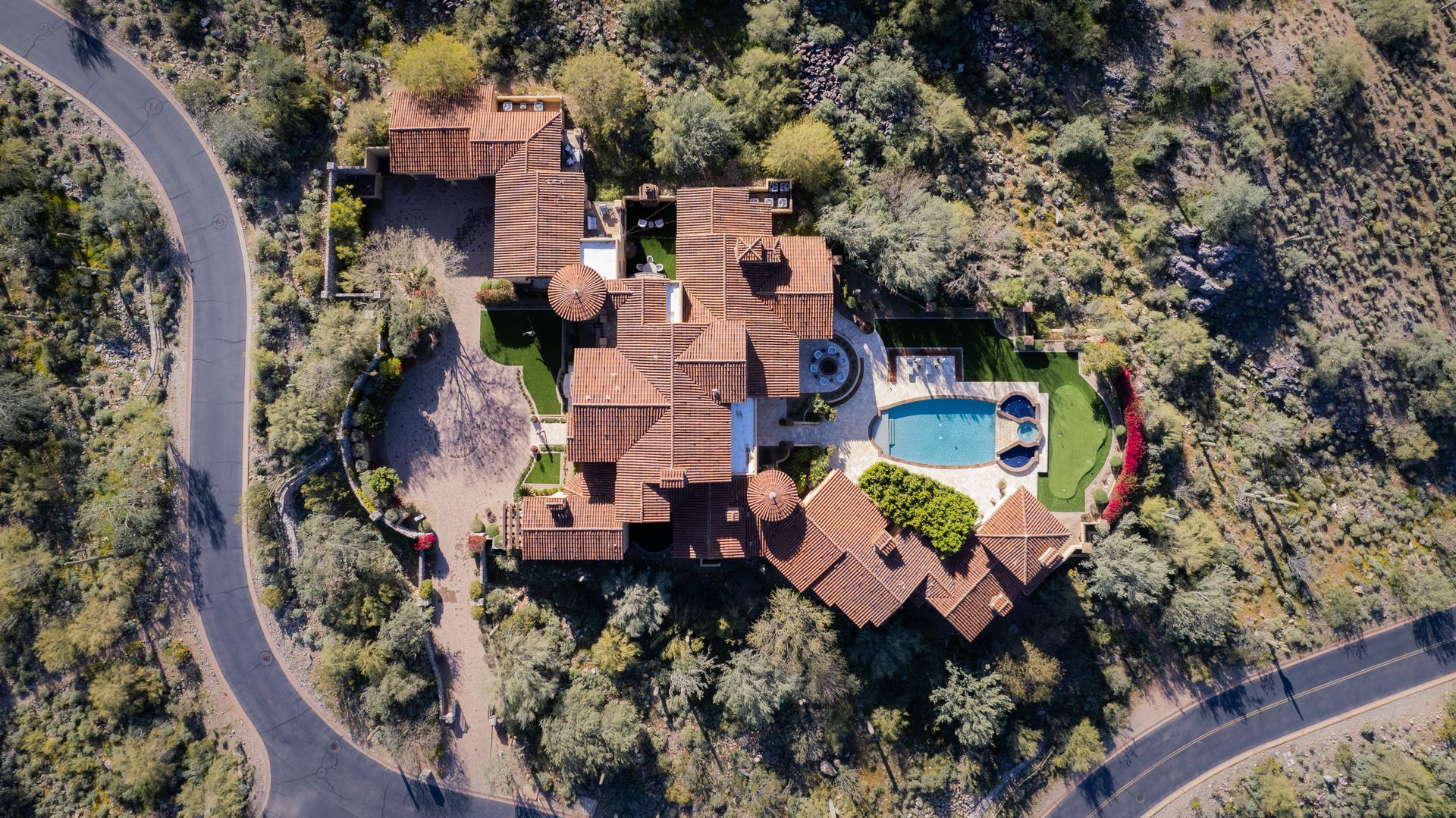 Aerial view of a large Spanish-style house with a pool, surrounded by trees and a winding road.