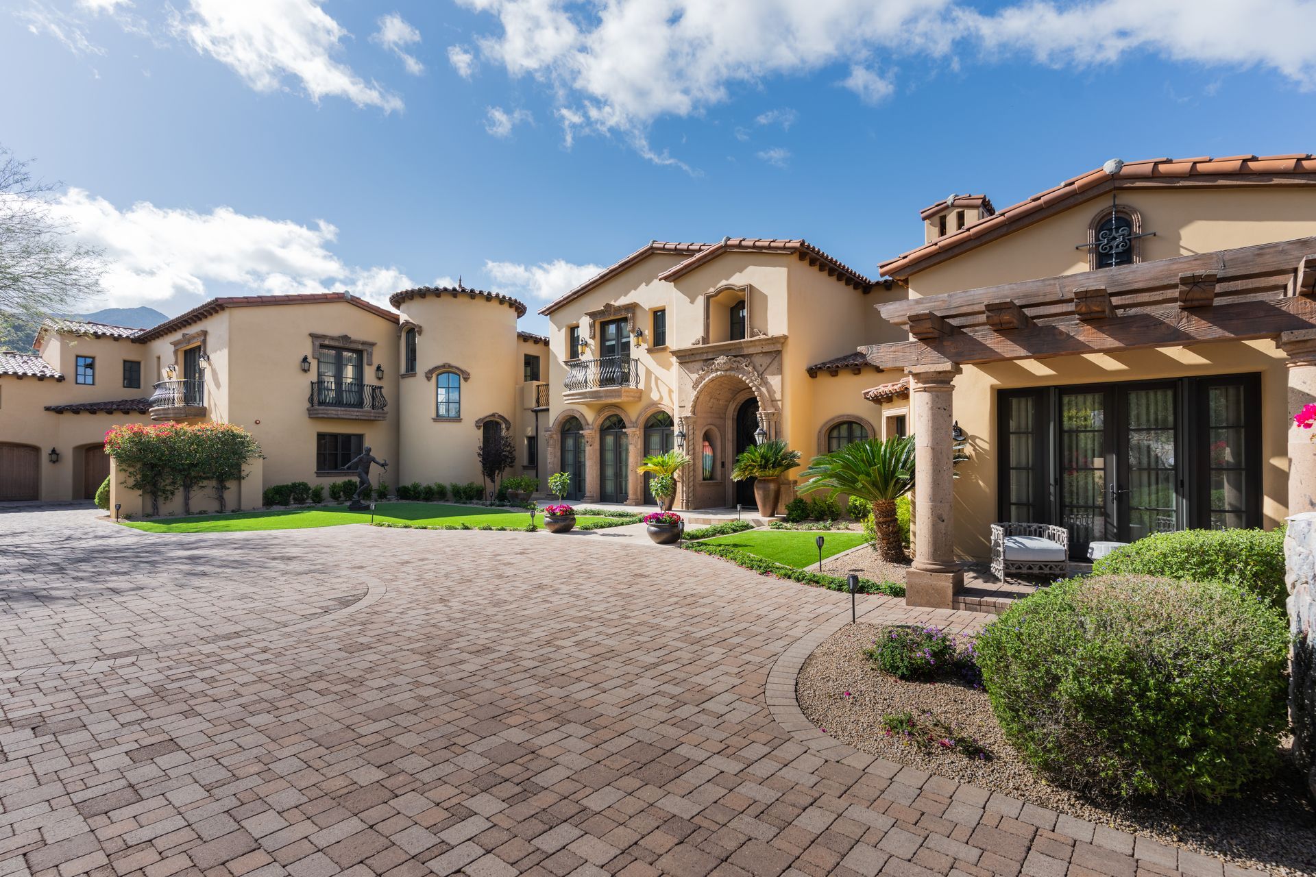 Large, ornate beige house with brick driveway, arched doorway, and blue sky.