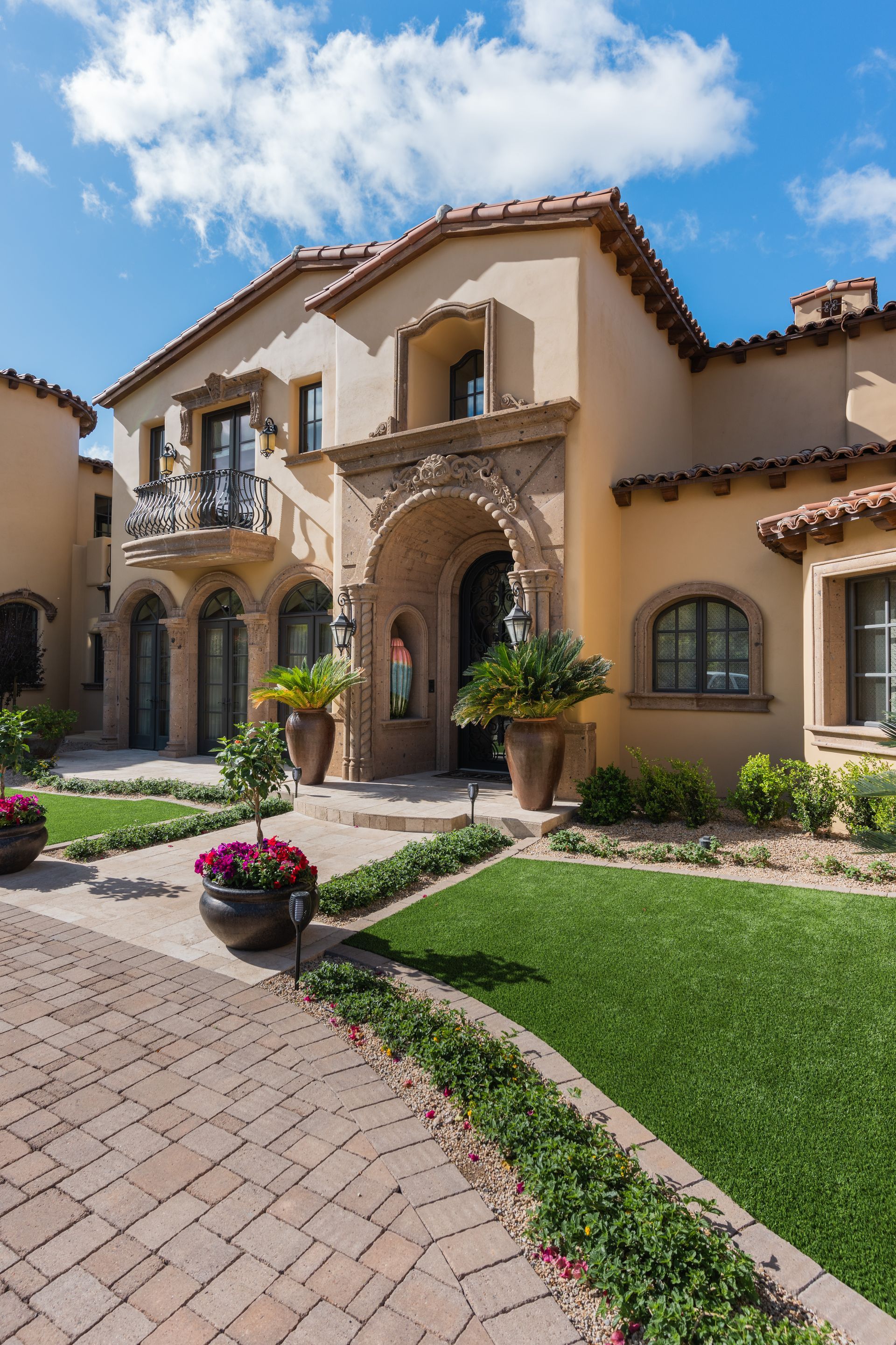 Beige Mediterranean-style house with arched entryway, brick pathway, green lawn, and blue sky.