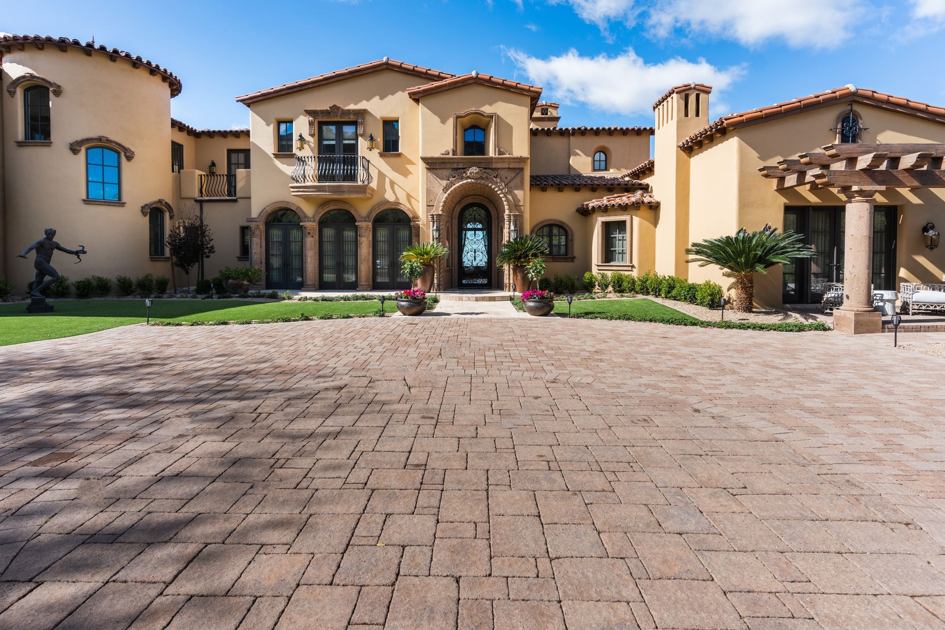 Beige stucco mansion with a brick driveway under a blue sky.