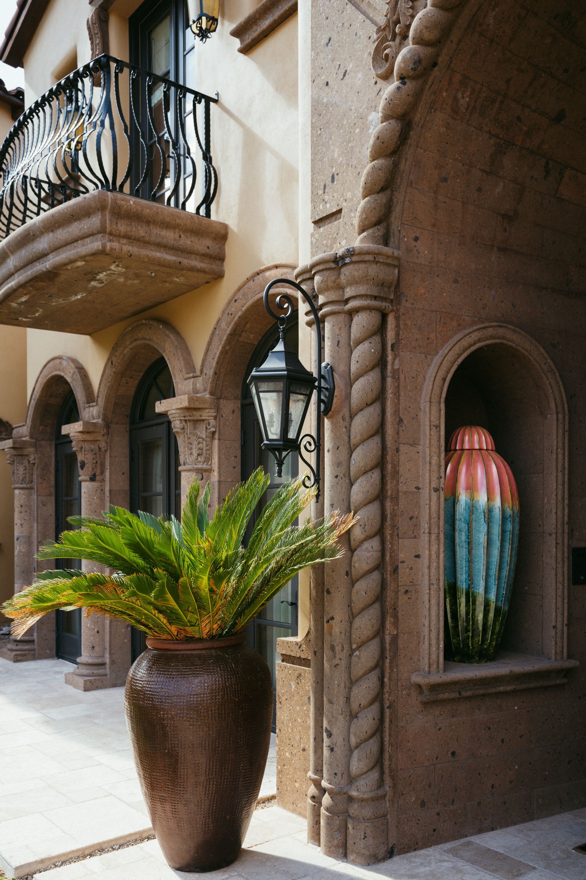 Exterior of a building with arched doorways and a balcony; large potted plant in the foreground.