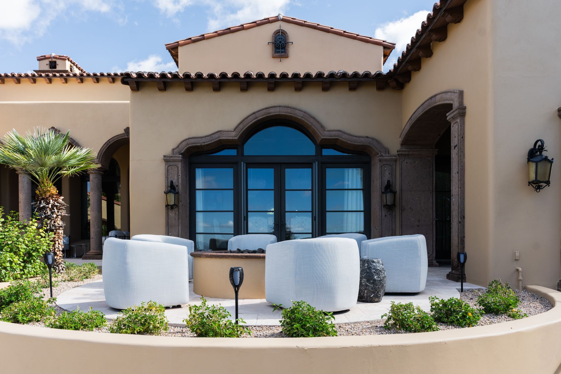 Patio seating area of a home; cream-colored stucco, arched window, white outdoor furniture, and small plants.