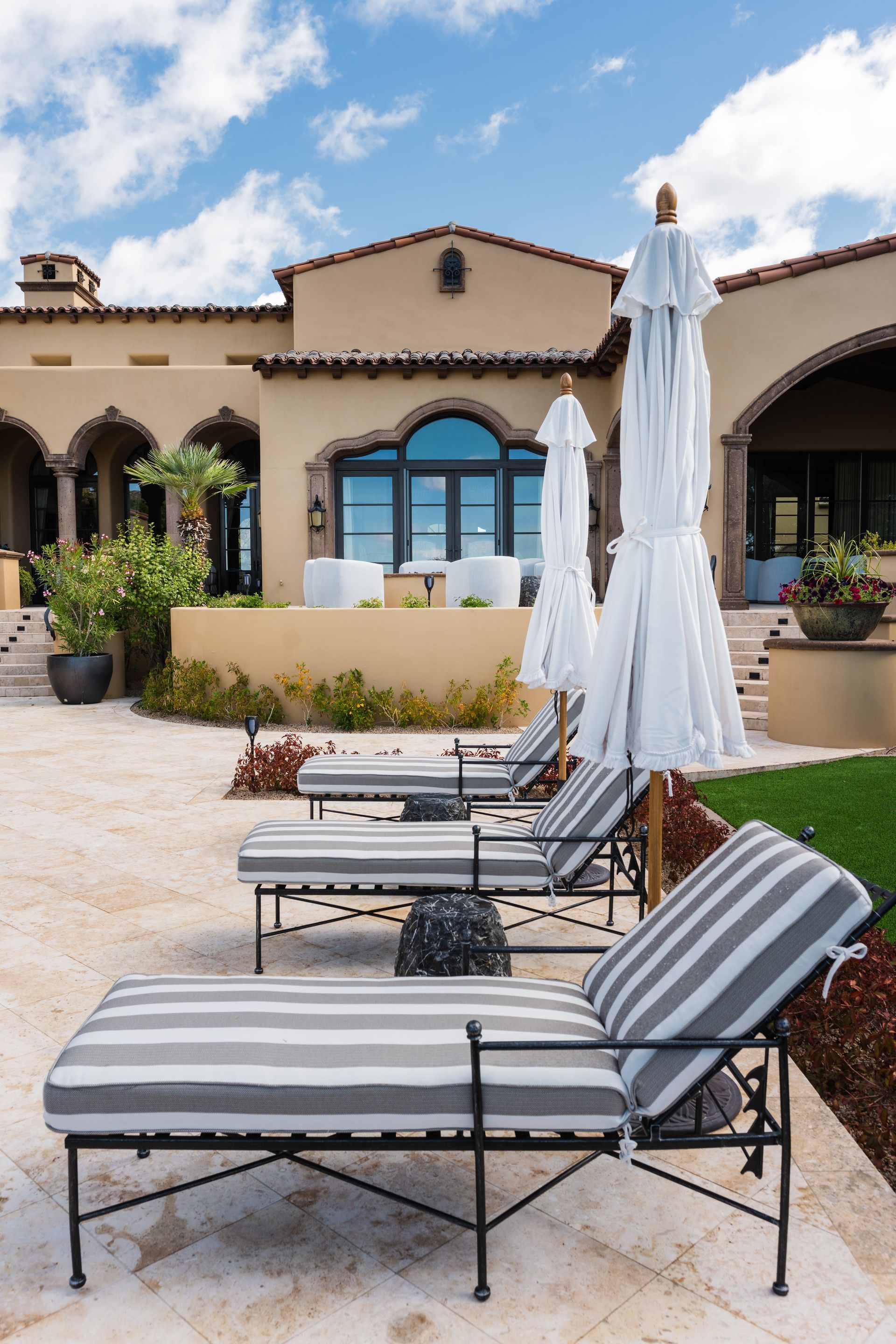 Lounge chairs with striped cushions and umbrellas on a patio by a stucco building, blue sky.